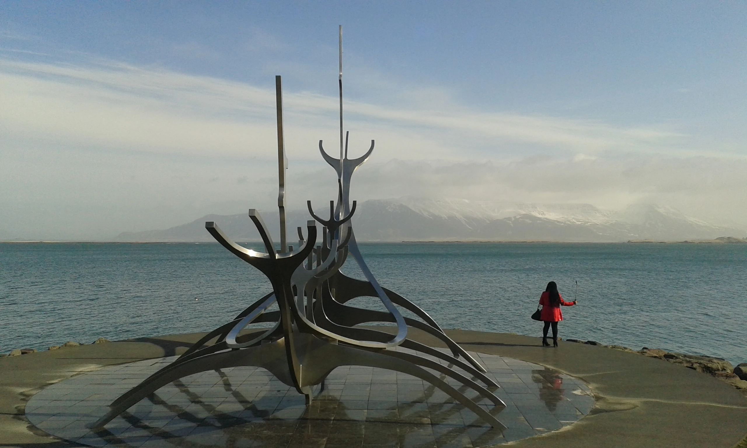 Island_Reykjavik Skulptur eines Wikingerbootes aus Metall am Ufer, im Hintergrund Meer und bewölkter Himmel, rechts eine Person mit rotem Mantel.