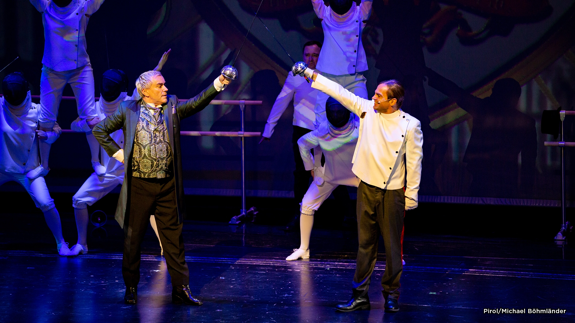 Musical Zeppelin: Zwei Männer auf Bühne in Theaterkostümen, einer im schwarzen Anzug mit gemustertem Hemd, der andere in weißer Jacke, beide heben die Fäuste zueinander