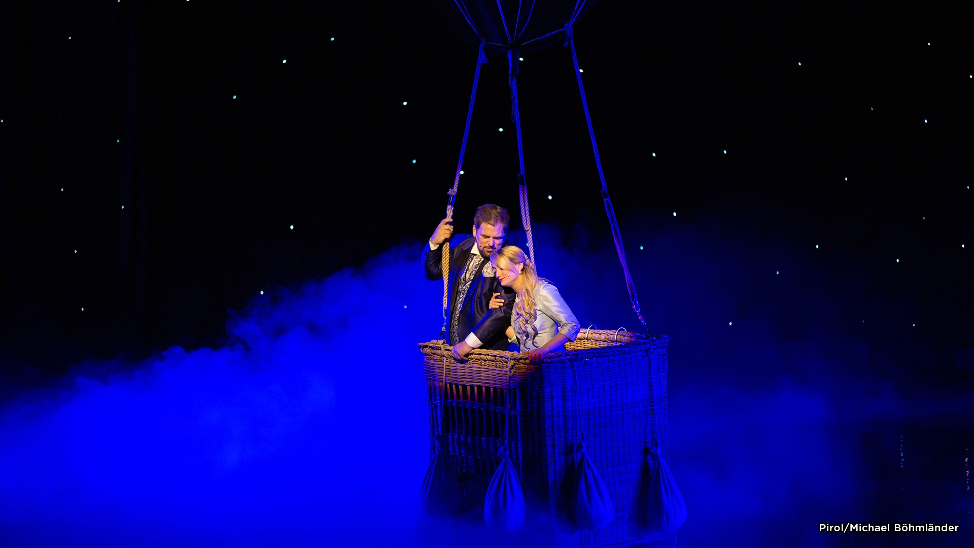 Musical Zeppelin: Zwei Personen stehen in einem Heißluftballonkorb, umgeben von blauem Nebel und einem dunklen Sternenhimmel