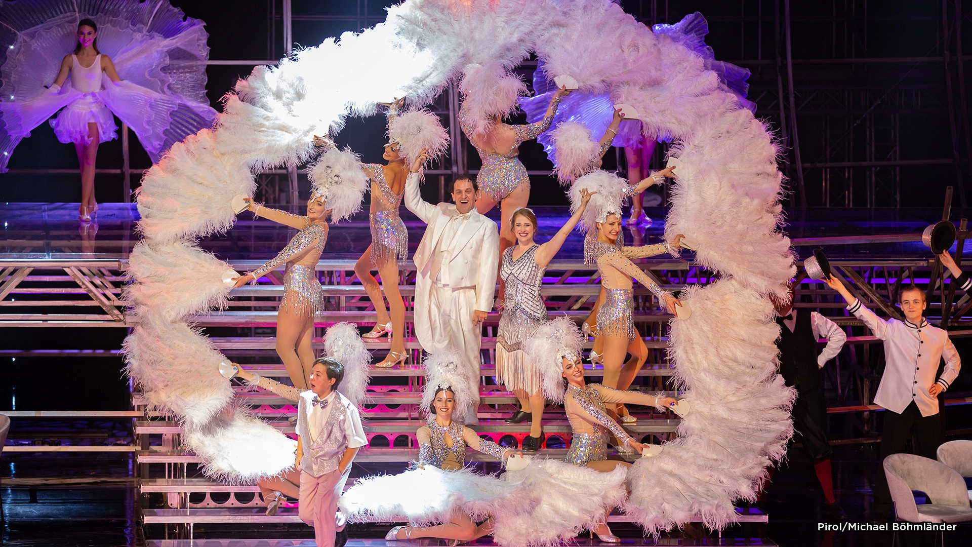 Musical Zeppelin: Tänzerinnen und Tänzer in glitzernden Kostümen mit großen weißen Federfächern auf einer Bühne, die eine kreisförmige Formation bilden.