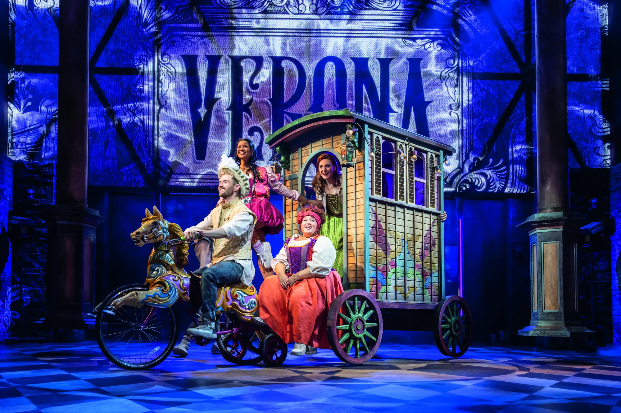 Eine farbenfrohe Theaterszene mit Darstellern auf einem skurrilen Wagen vor einem leuchtend blauen Hintergrund, auf dem das Wort „VERONA“ steht.