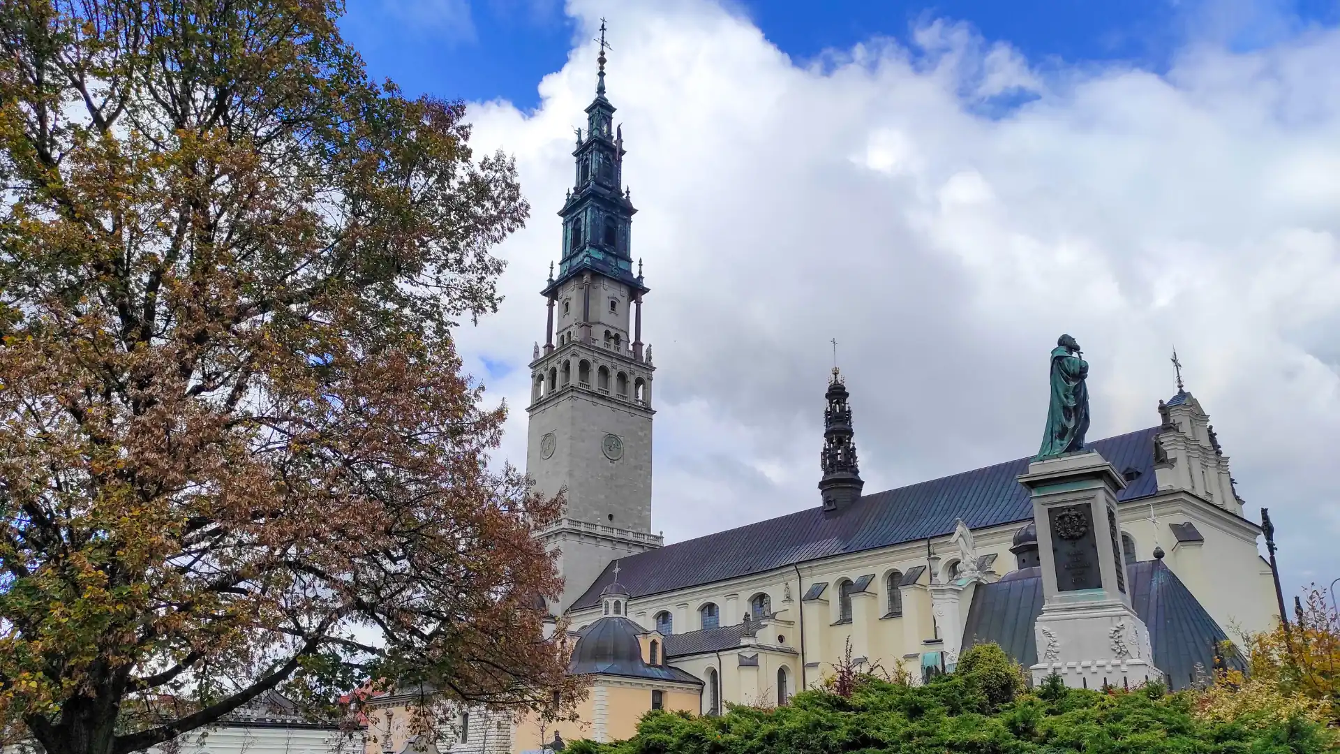 Die Basilika des Heiligen Antonius von Padua in Częstochowa mit einem hohen Turm und einer Statue im Vordergrund, umgeben von Bäumen und bewölktem Himmel