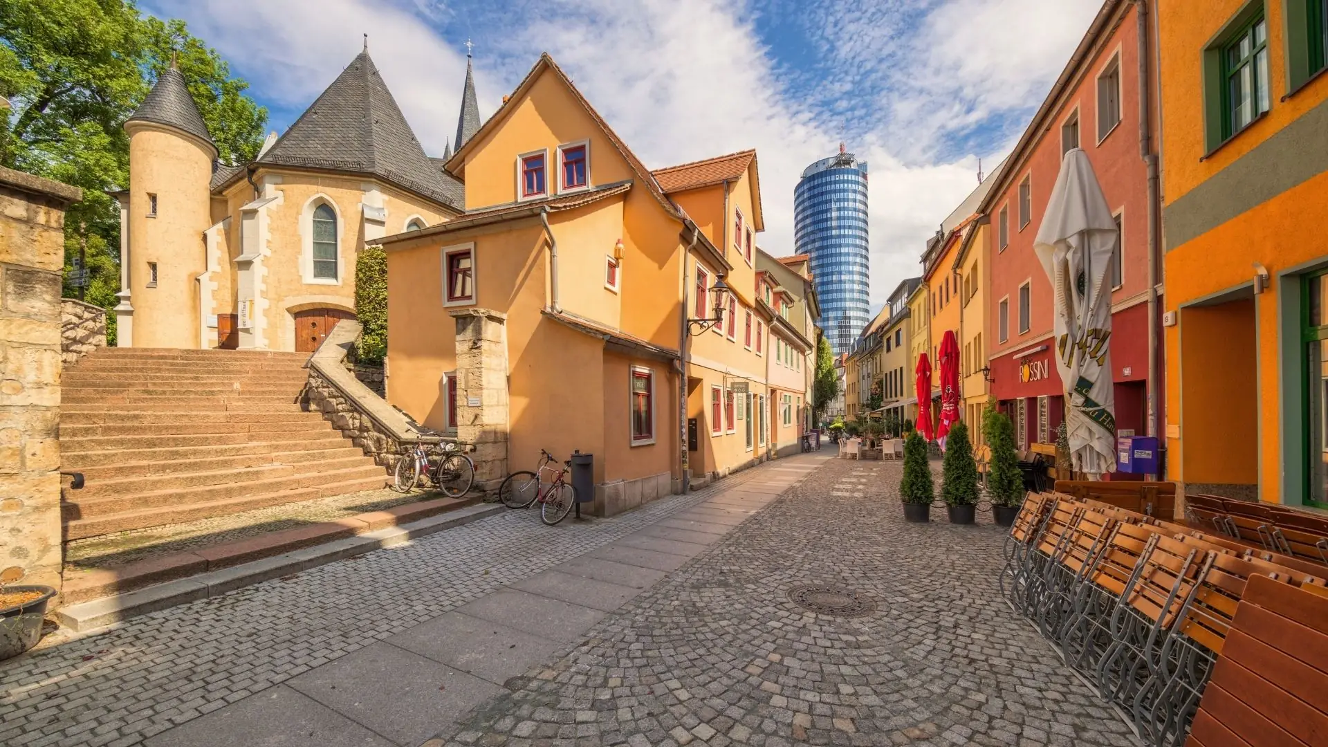 Blick auf eine gepflasterte Straße in der Altstadt von Jena, mit bunten Gebäuden, einer Treppe und einem modernen Hochhaus im Hintergrund.