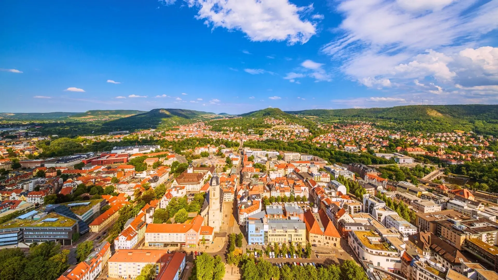 Luftaufnahme von Jena mit roten Dächern, grünen Bäumen und Hügeln im Hintergrund unter blauem Himmel mit Wolken.