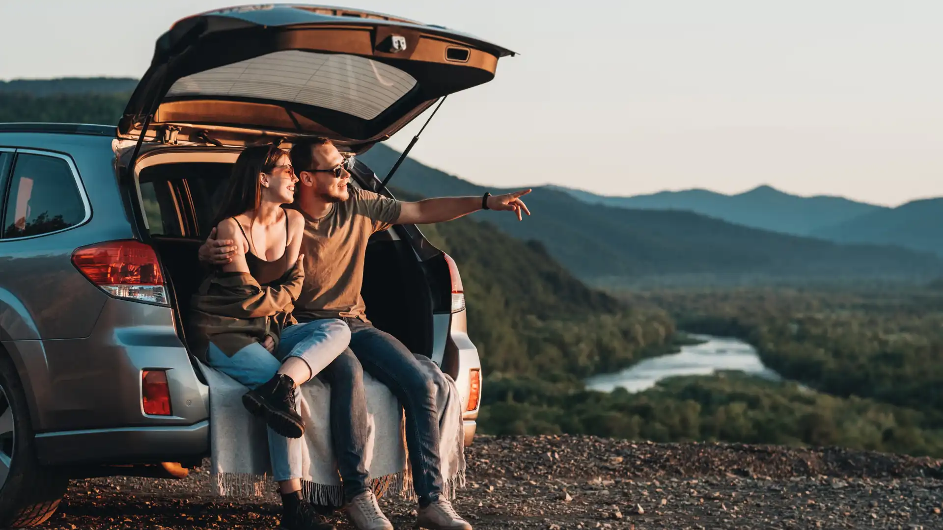 Junges Paar auf Roadtrip sitzt im geöffneten Kofferraum ihres Autos während des Sonnenuntergangs.