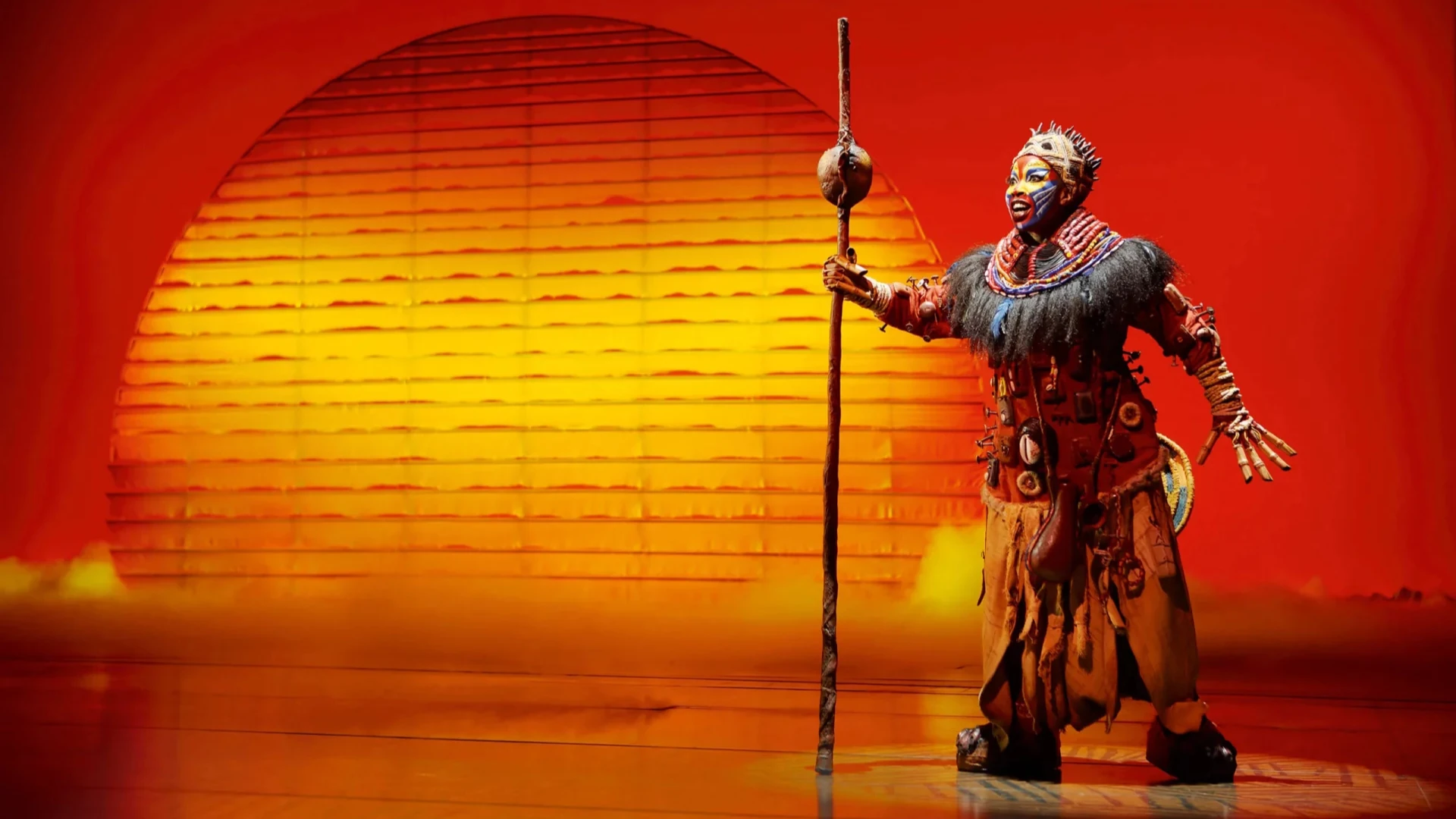 Ein Darsteller in aufwändiger Stammeskleidung, der einen Stab in der Hand hält, steht vor einem leuchtenden Sonnenuntergangshintergrund in Orange- und Gelbtönen.