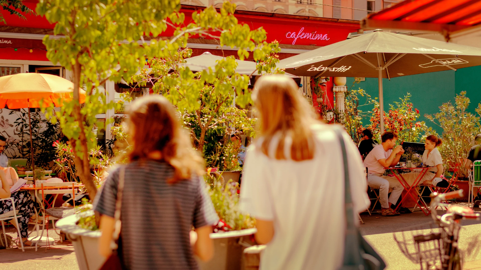 Dieses Foto zeigt die lebendige Atmosphäre am Karmelitermarkt in Wien. Im Vordergrund schlendern zwei Personen durch die belebte Marktszene, während Menschen unter Sonnenschirmen in gemütlichen Cafés sitzen. Die bunten Fassaden, üppige Pflanzen und warme Lichtstimmung verleihen dem Ort einen charmanten und einladenden Charakter.