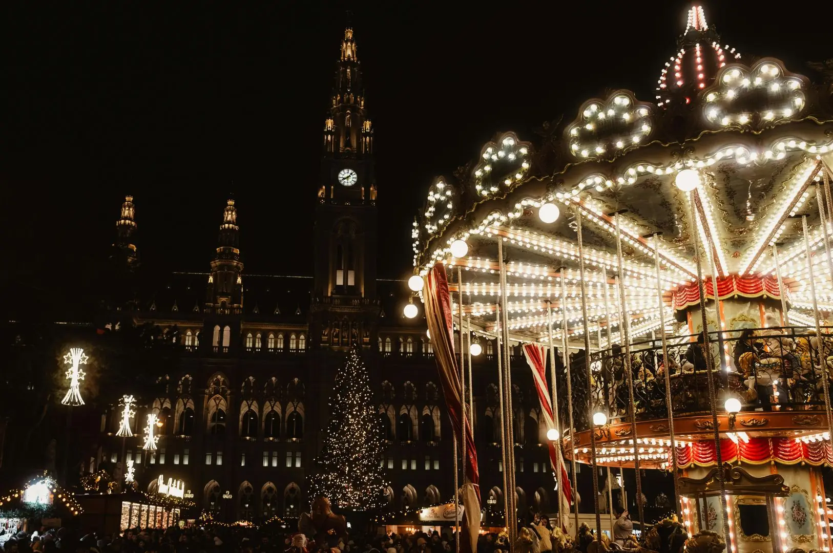 Beleuchtetes Karussell im Vordergrund, im Hintergrund das Rathaus von Wien mit Uhr und Weihnachtsbaum. Nachtaufnahme.