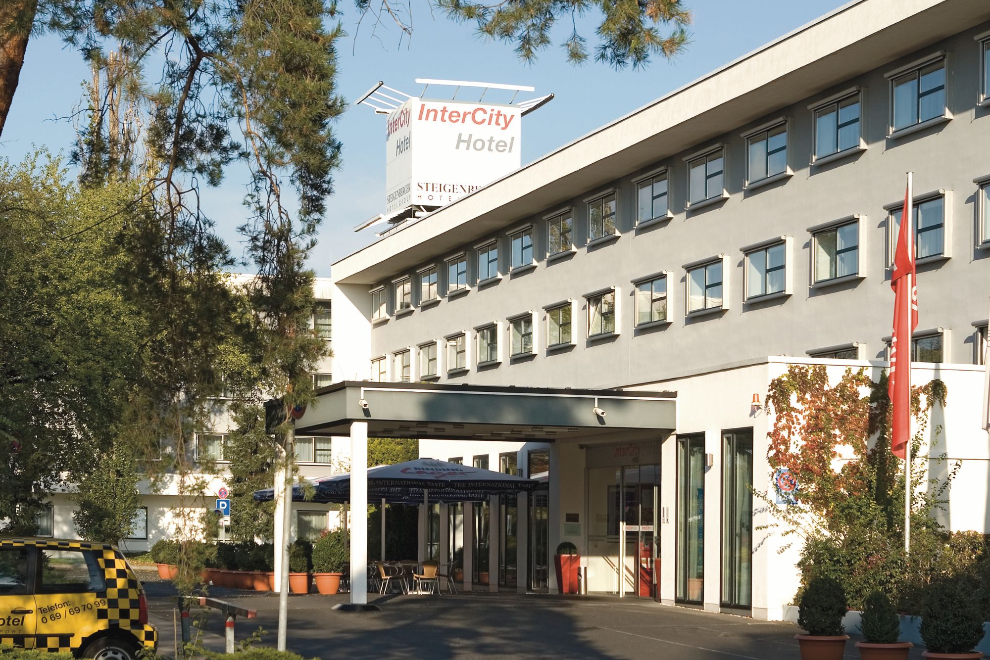 Außenansicht des InterCity Hotels mit modernem Design, Eingangsmarkise und einem Taxi, das in der Nähe einer Grünanlage wartet.