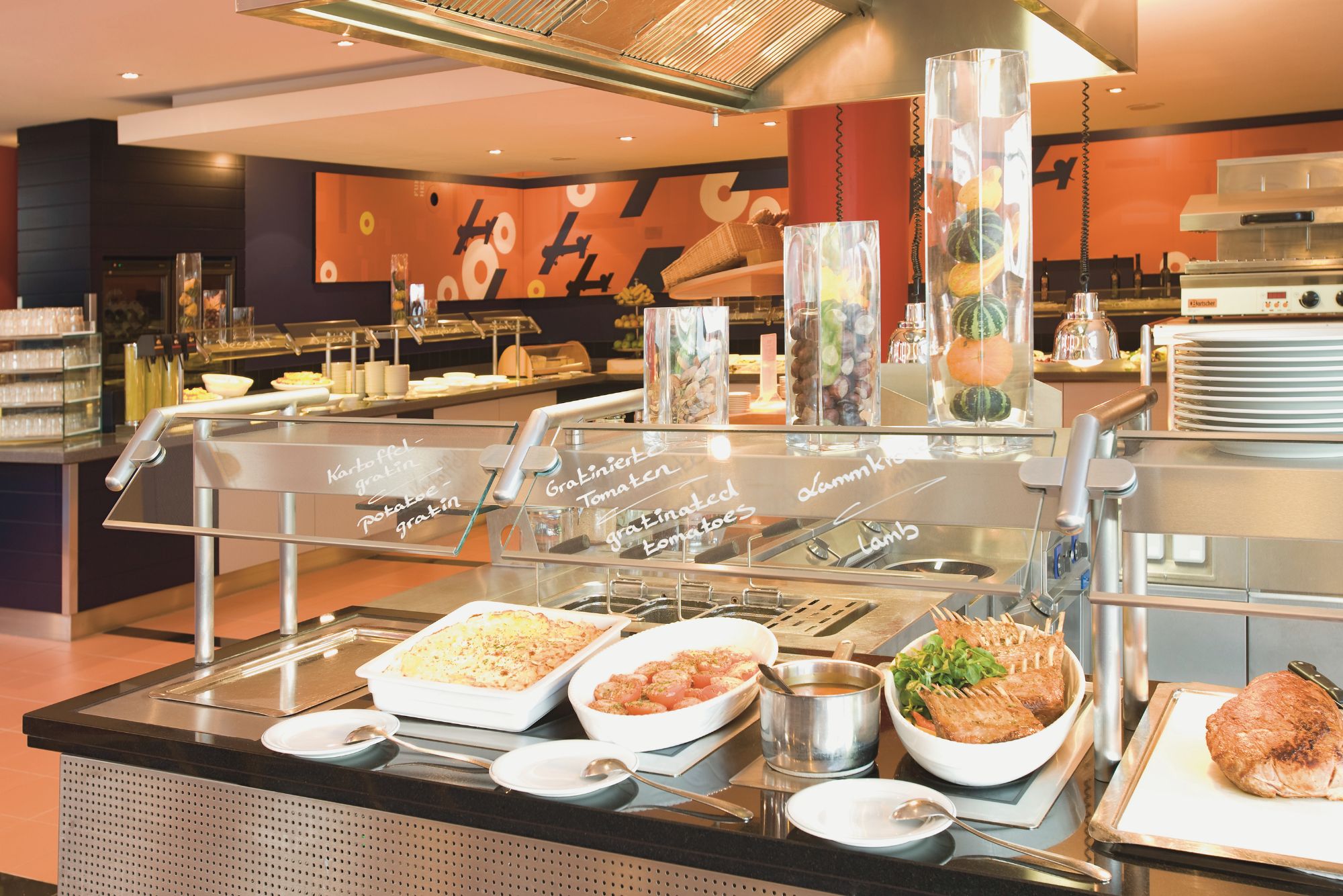 Ein modernes Buffet mit verschiedenen Gerichten wie Kartoffelgratin, Lammfleisch und Salat, mit bunten Dekorationen im Hintergrund.