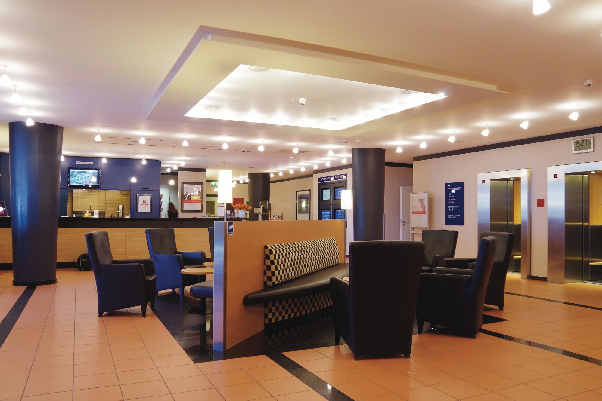 Eine moderne Hotellobby mit dunklen Ledersesseln, einem Empfangstresen, einer beleuchteten Decke und Aufzügen im Hintergrund. Warme, einladende Atmosphäre.