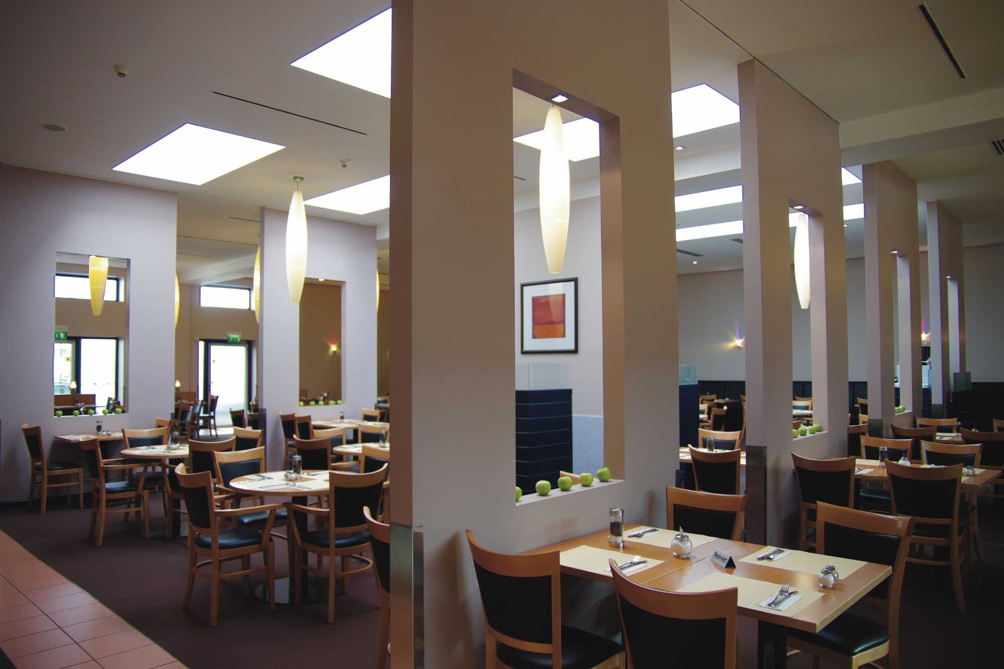 Modernes Restaurantinterieur mit Holztischen, Stühlen und hellen Oberlichtern, mit grünen Äpfeln entlang einer Trennwand.