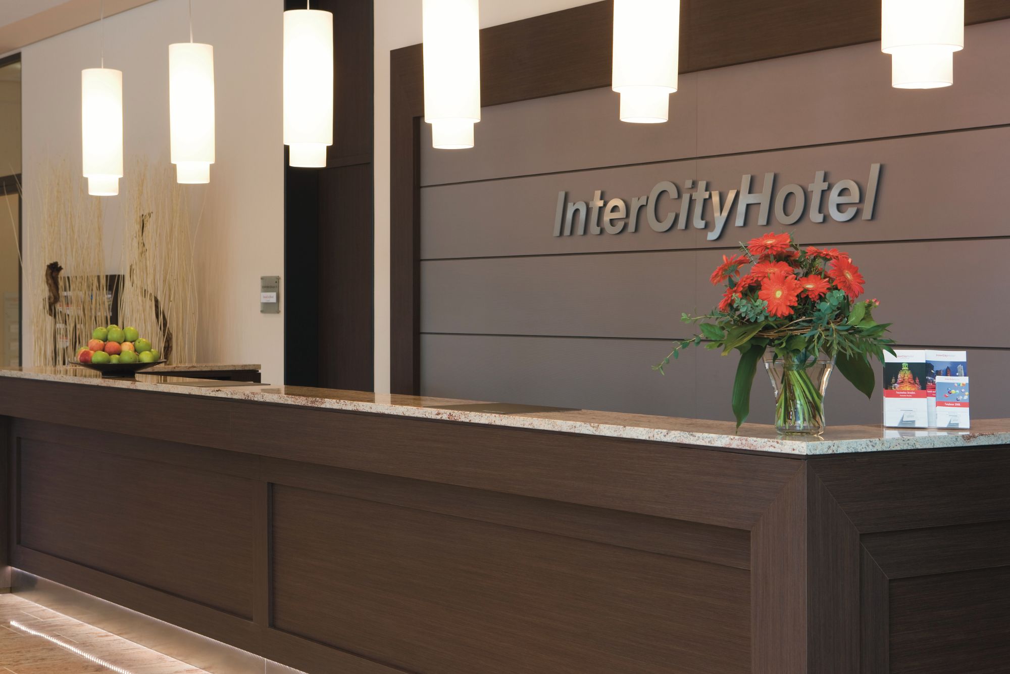 Eine moderne Hotelrezeption mit einem Logo, frischen Blumen und einer Schale mit buntem Obst. Eine elegante Beleuchtung unterstreicht die einladende Atmosphäre.