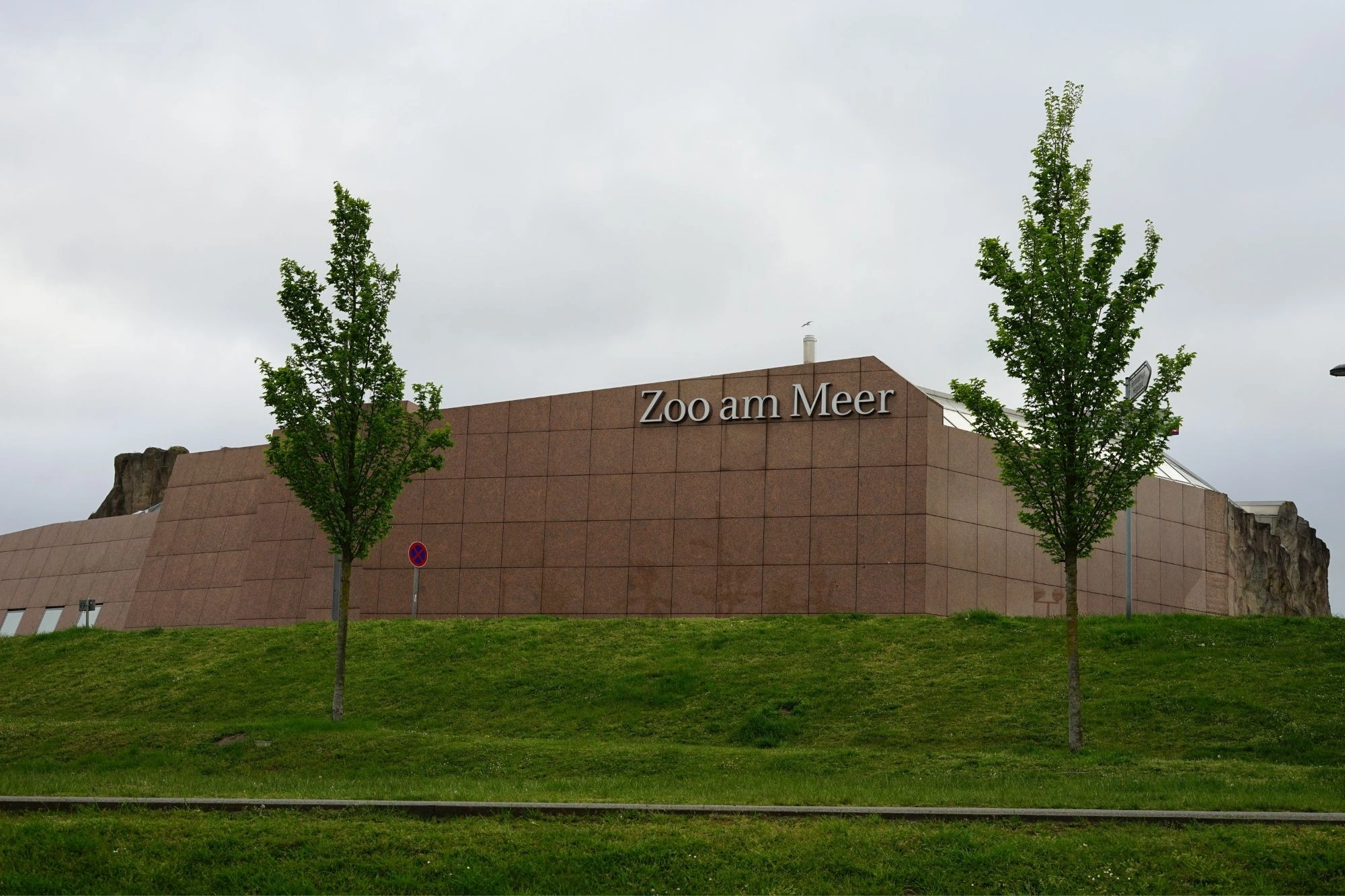 Eingang des Zoos am Meer mit braunem Gebäude und zwei Bäumen im Vordergrund