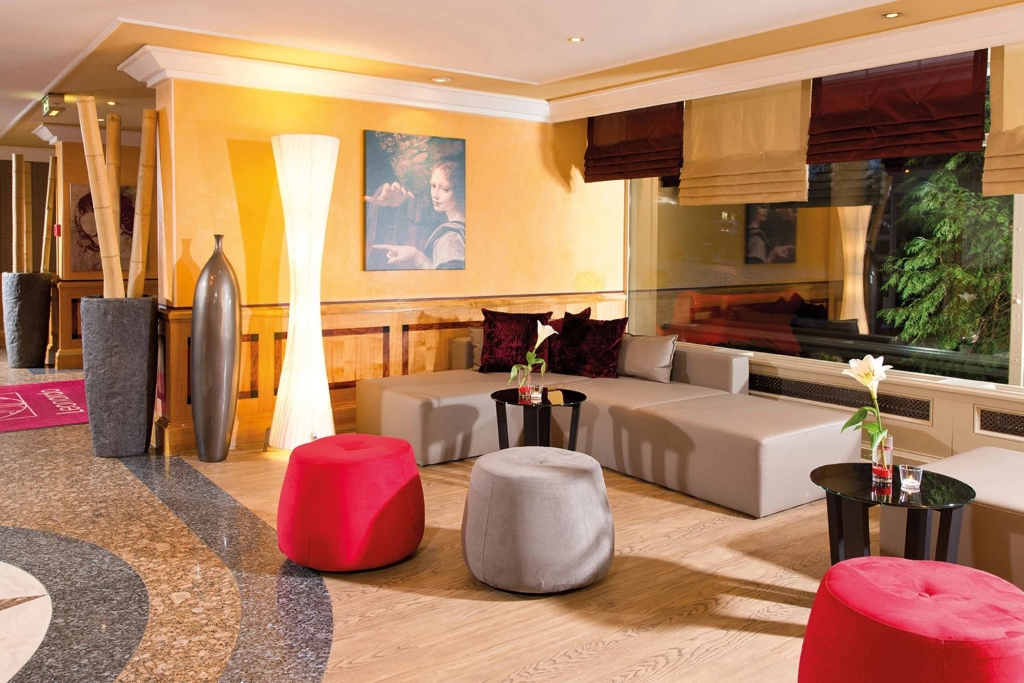 Gemütliche Hotellobby mit grauer Eckcouch, roten und grauen Sitzhockern, kleinen schwarzen Tischen mit Blumen und großen Vasen neben einer Stehlampe