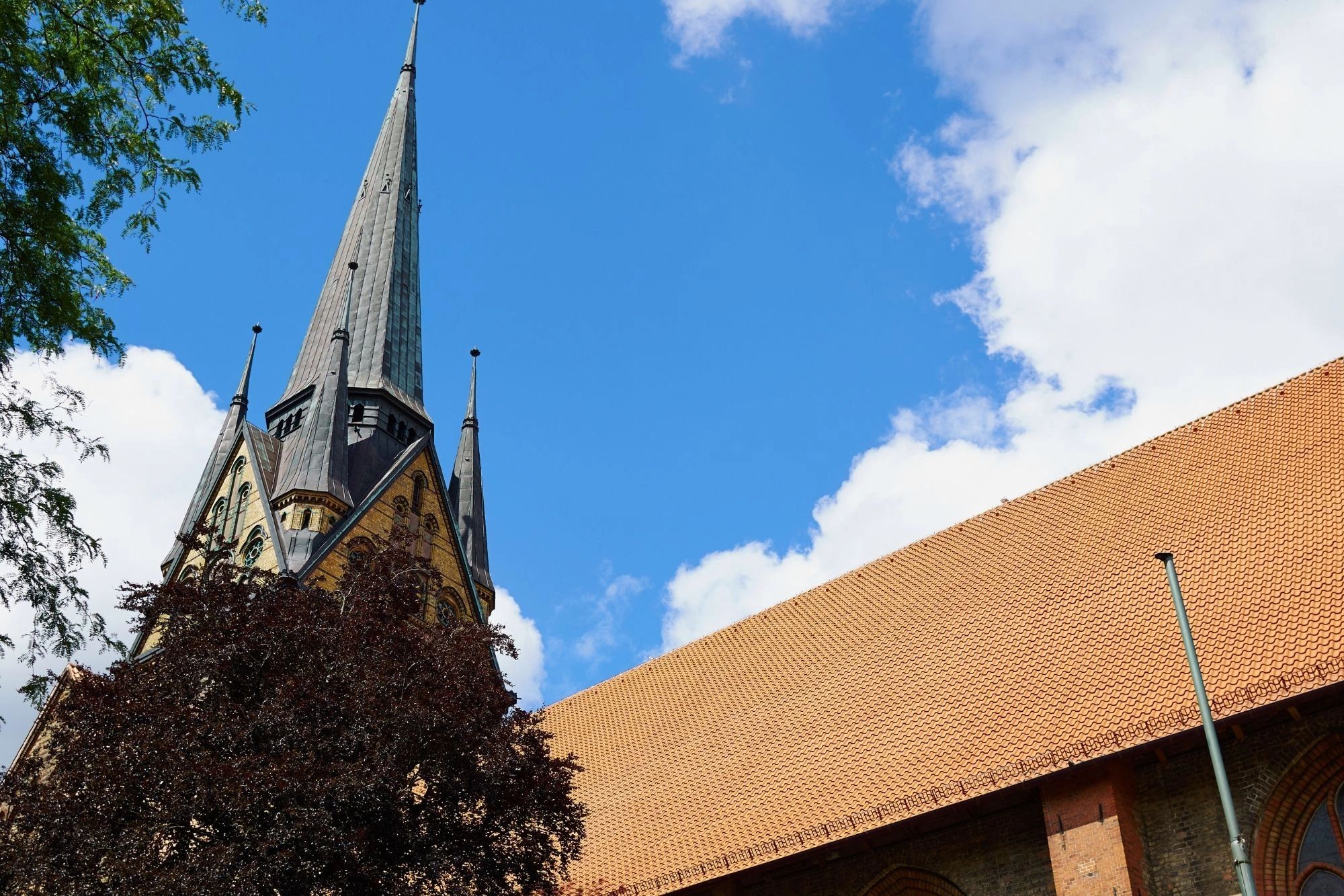 Kirchturm mit spitzem Dach und zwei kleineren Türmchen, davor ein Baum, blauer Himmel mit Wolken.