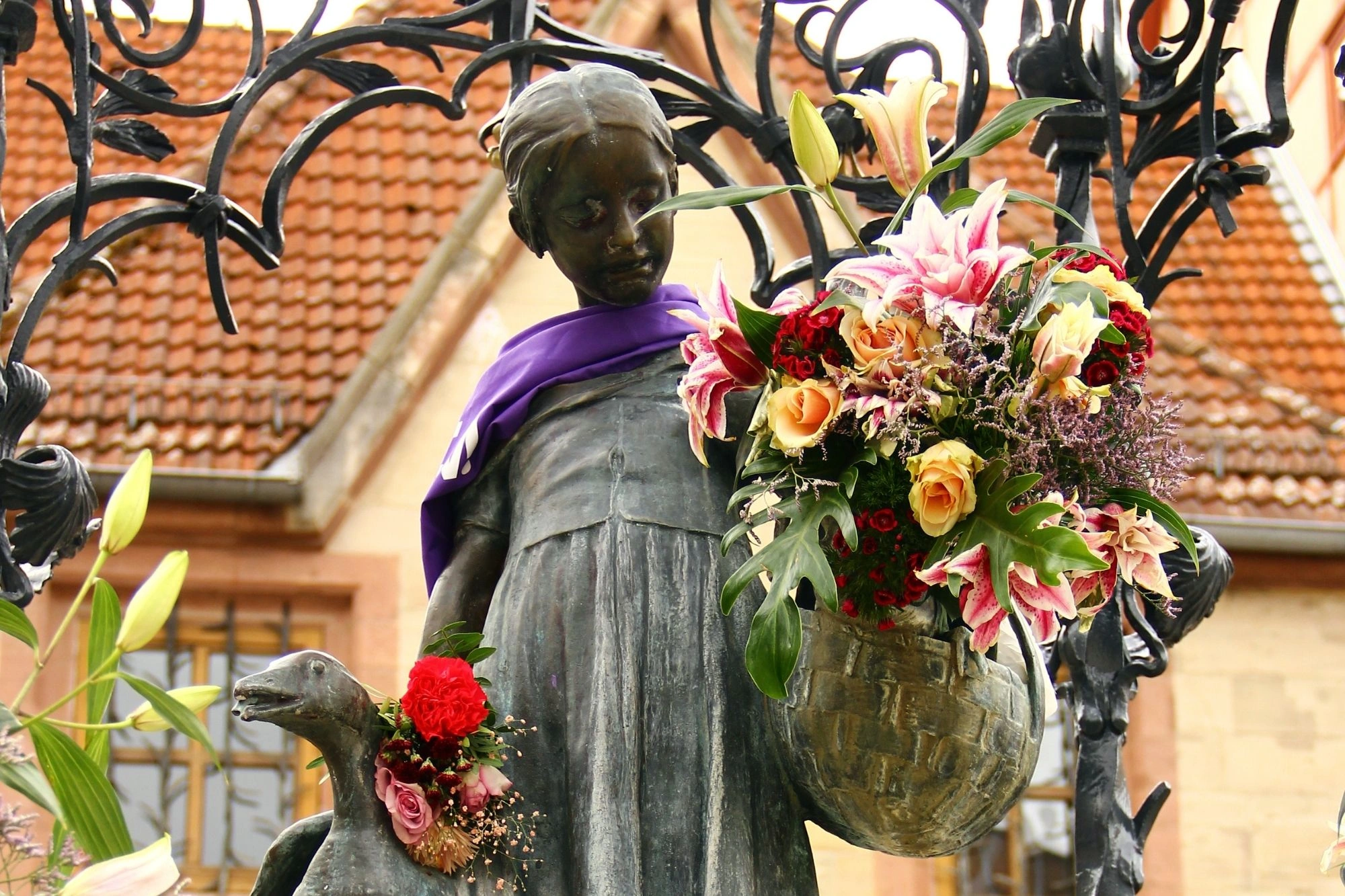 Bronzestatue einer stehenden Person mit violettem Schal, umgeben von Blumen und einem Vogel mit Blumenschmuck am Hals.