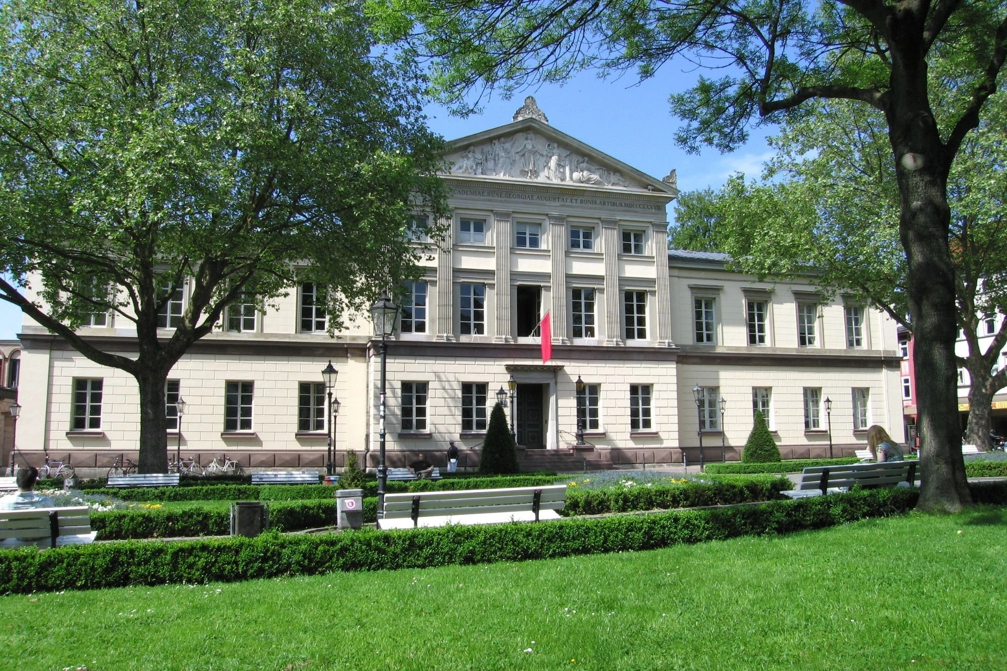 Neoklassizistisches Gebäude mit Säulen und Dreiecksgiebel, davor gepflegter Park mit Bänken und Büschen, zwei große Bäume rahmen die Szene.