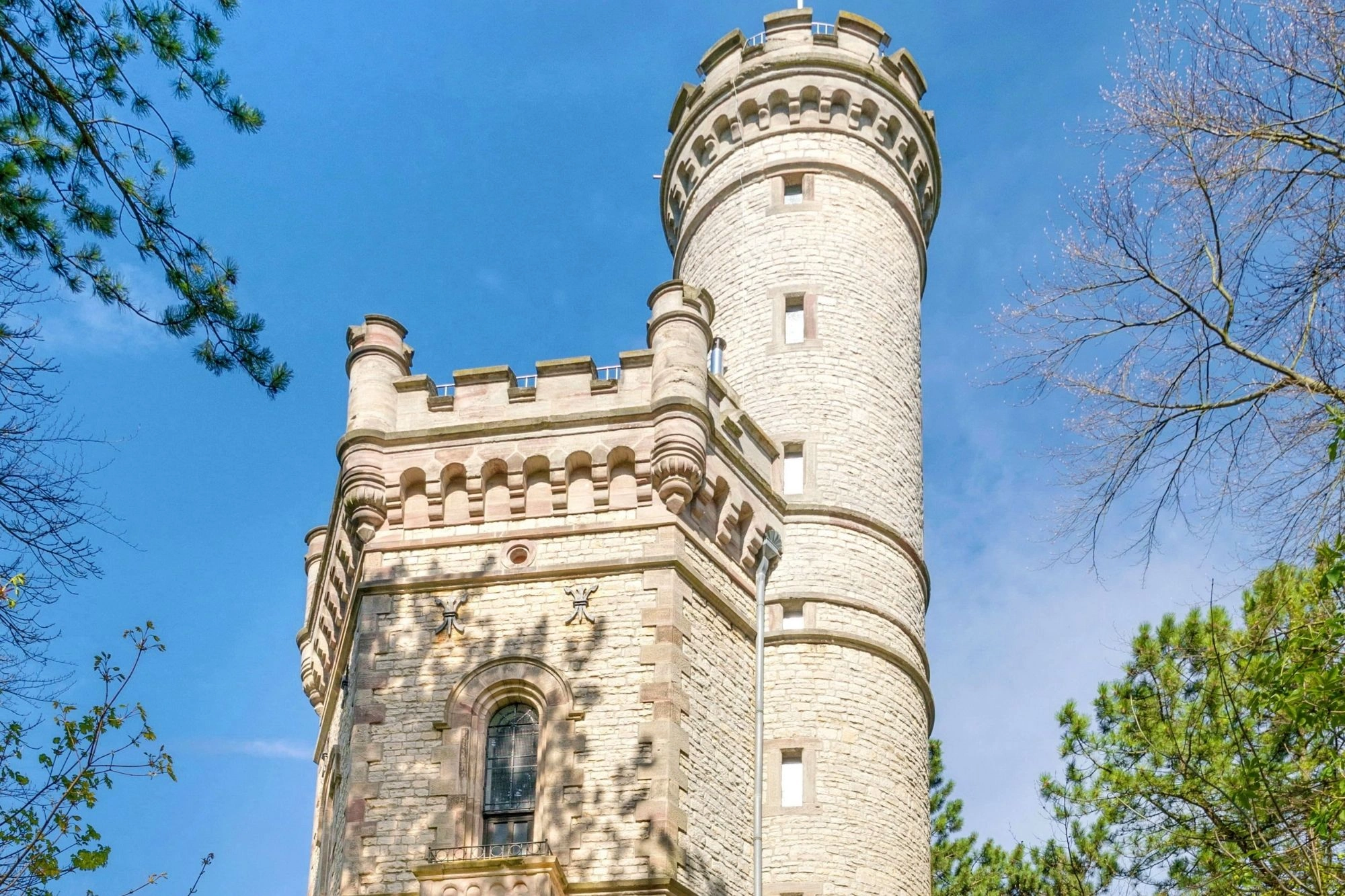 Turm aus hellem Stein mit Zinnen und Rundturm vor blauem Himmel und Bäumen.
