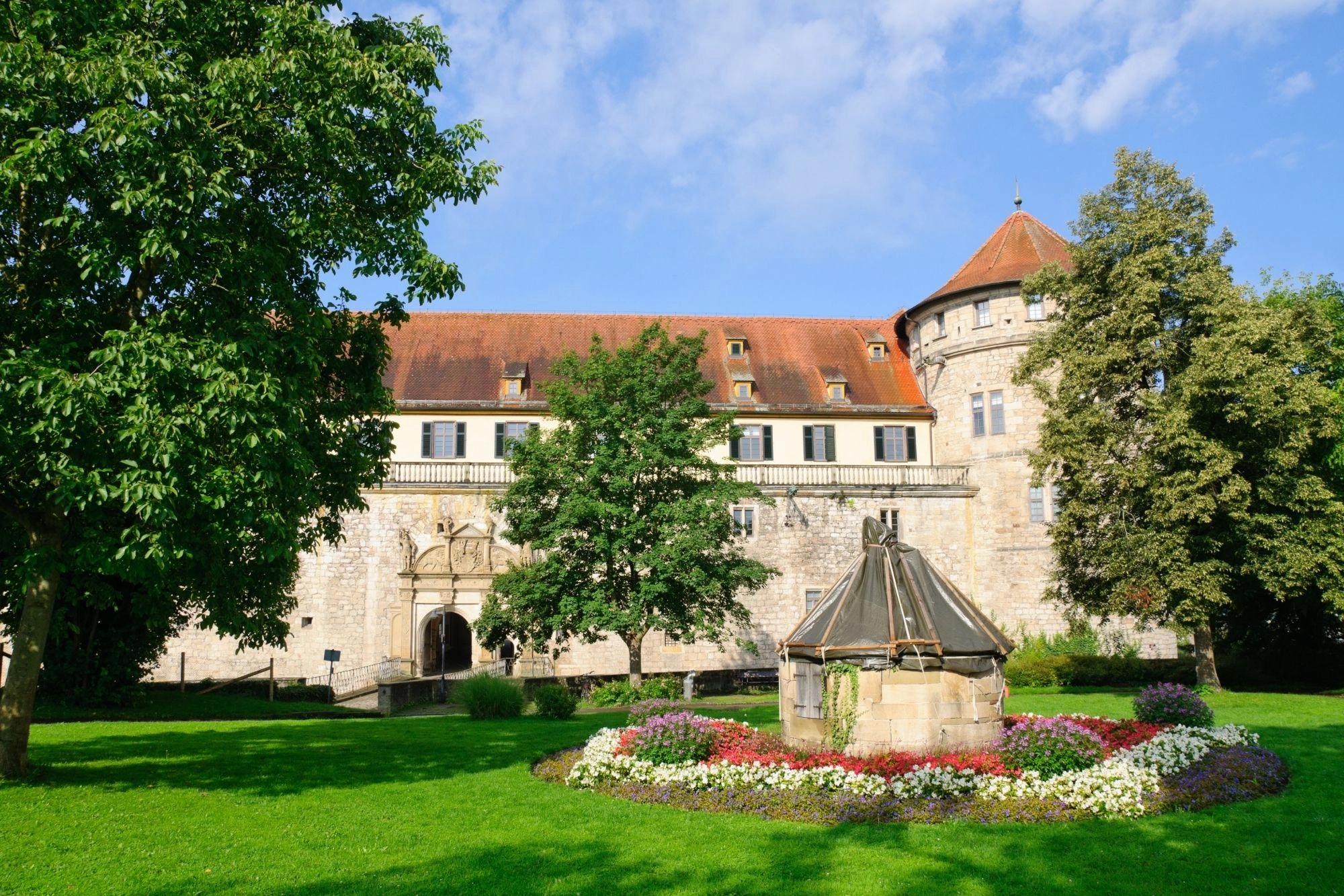 Historisches Schloss mit rotem Ziegeldach, Rundturm rechts, umgeben von grünen Bäumen und einem Blumenbeet mit Brunnen im Vordergrund.