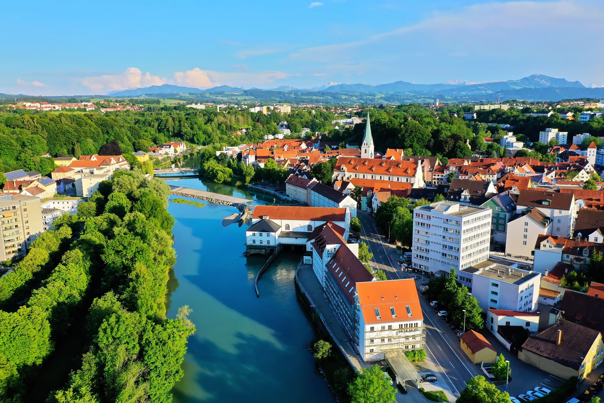 Das Bild zeigt einen Luftblick auf die Stadt Kempten im Allgäu, eingebettet in eine grüne Landschaft. Im Vordergrund fließt ein ruhiger Fluss, der von malerischen Häusern mit roten Dächern gesäumt wird. Die historische Architektur der Altstadt und moderne Gebäude fügen sich harmonisch in das Stadtbild ein. Im Hintergrund ragen die imposanten Berge des Allgäus empor und bilden einen eindrucksvollen Kontrast zur lebendigen Stadt. Das Bild vermittelt eine ruhige, aber dennoch dynamische Atmosphäre, typisch für Kempten.