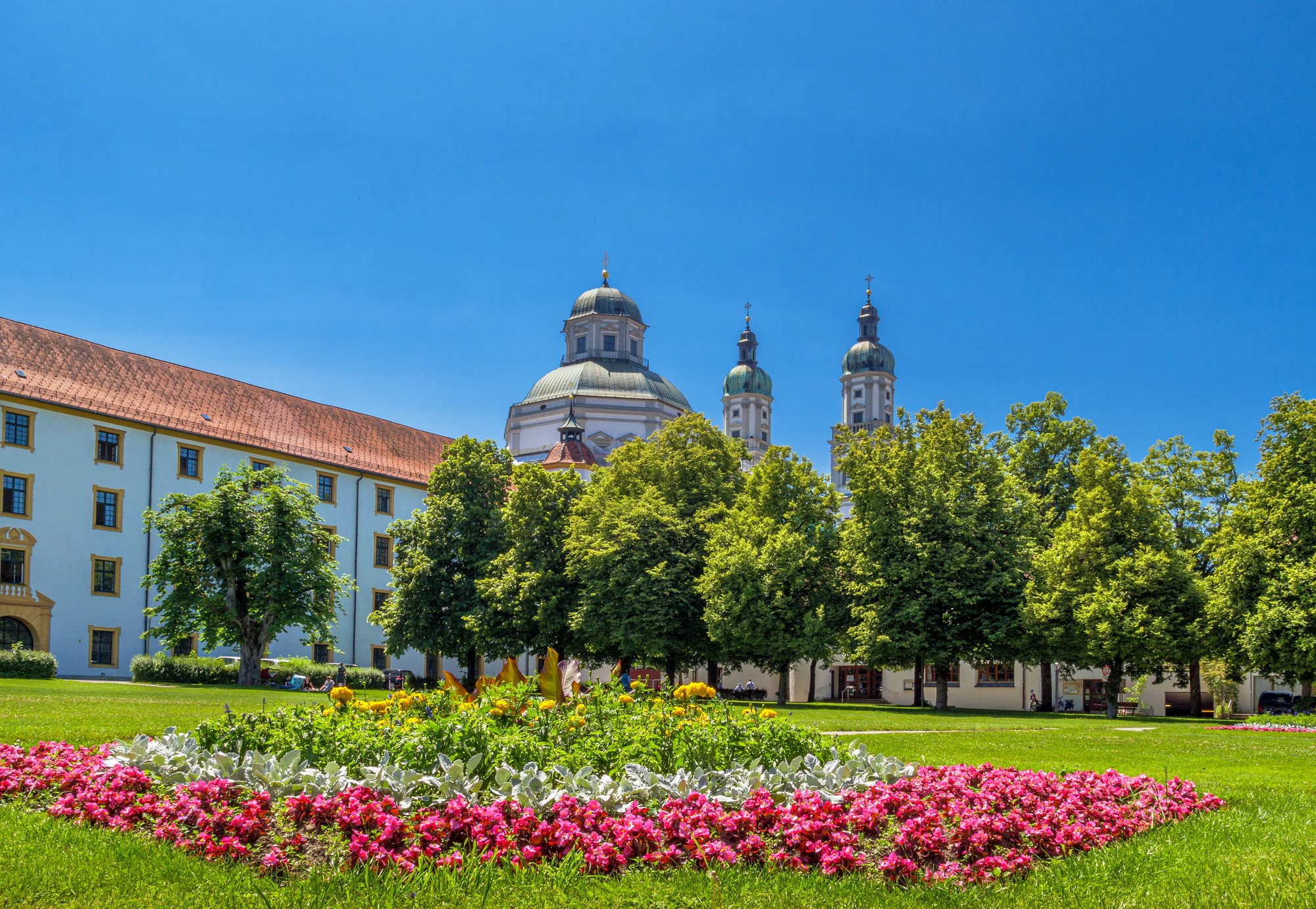 Das Bild zeigt den malerischen Garten des Kemptener Stiftungsareals, mit dem beeindruckenden barocken Gebäude im Hintergrund. Der weitläufige Rasen ist von grünen Bäumen umgeben und durch bunte Blumenbeete, darunter leuchtend rosa und gelbe Blumen, verschönert. Die markanten Zwiebeltürme der St. Lorenz Basilika ragen majestätisch in den strahlend blauen Himmel und verleihen der Szene eine historische Note. Das Bild vermittelt eine ruhige und einladende Atmosphäre, typisch für Kempten.