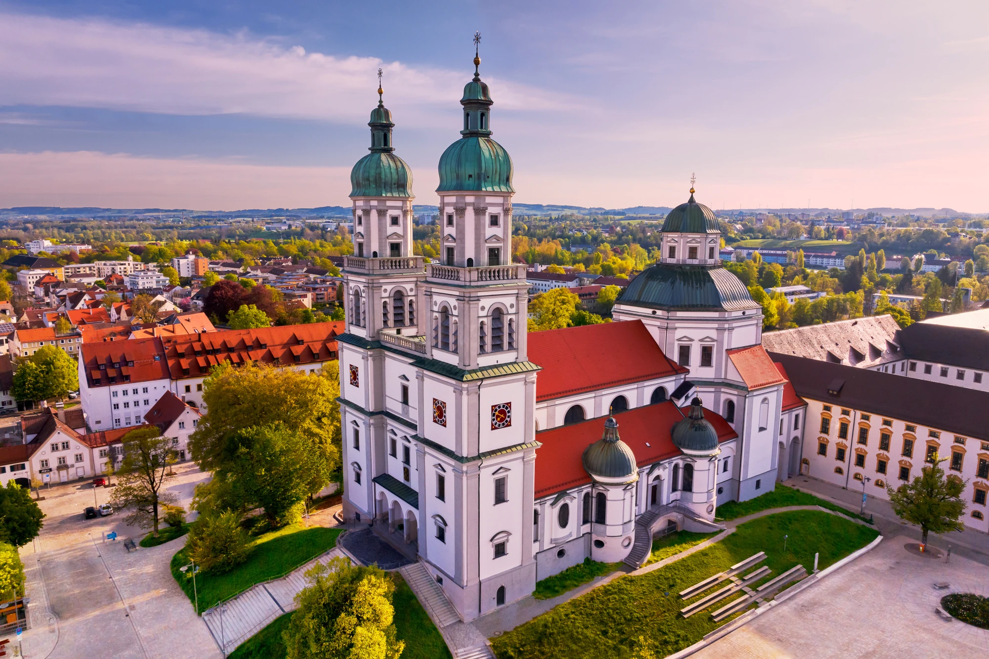 Das Bild zeigt die imposante Basilika St. Lorenz in Kempten, ein Meisterwerk barocker Architektur. Mit ihren markanten Zwiebeltürmen und dem prächtigen Kuppeldach ragt die Kirche über die umliegenden Gebäude der Stadt. Im Vordergrund sind die weitläufigen Grünflächen und der Platz gut zu erkennen, die das Bauwerk umgeben. Die Sonne taucht die Szene in warmes Licht und hebt die Farben der Stadt und der Natur hervor. Die Weite der Landschaft im Hintergrund bietet einen eindrucksvollen Blick auf die Umgebung von Kempten.