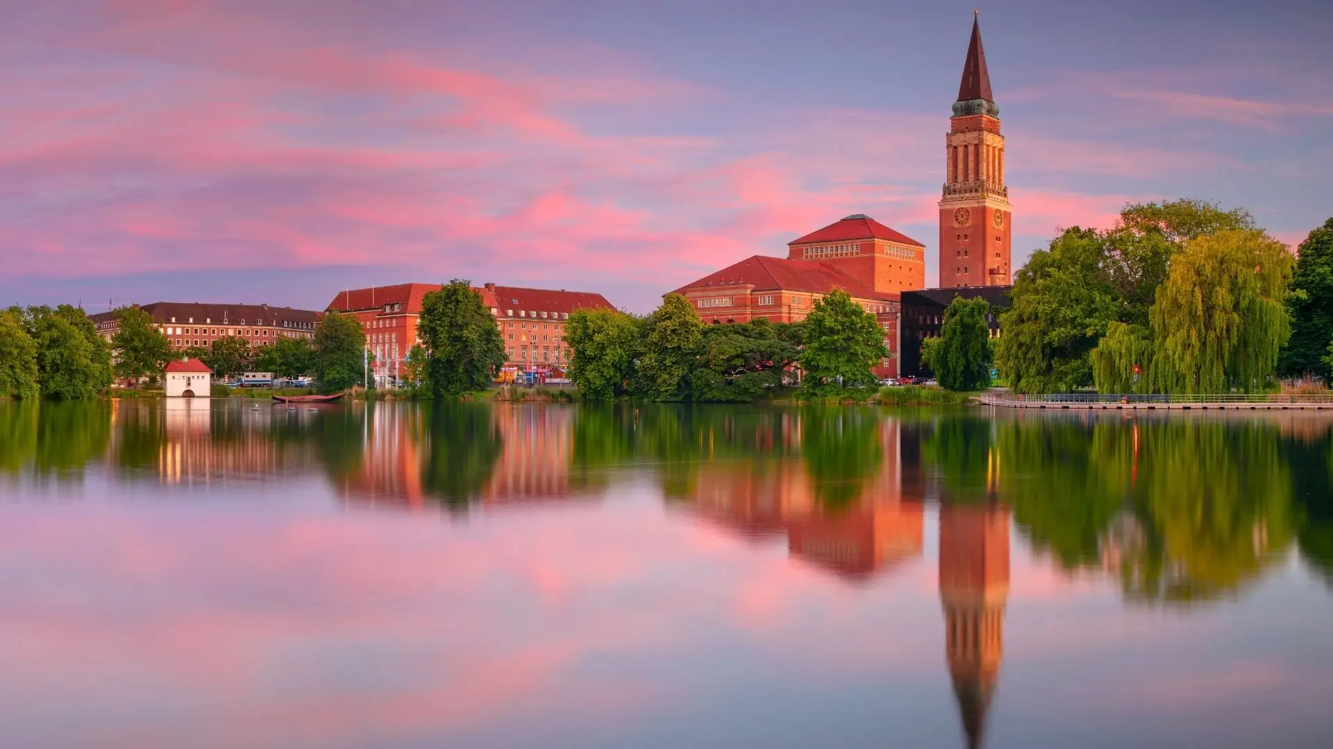 Panoramablick auf Kiel mit dem historischen Gebäude der Stadtverwaltung und dem Kirchturm im Hintergrund, reflektiert im ruhigen Wasser des Sees.