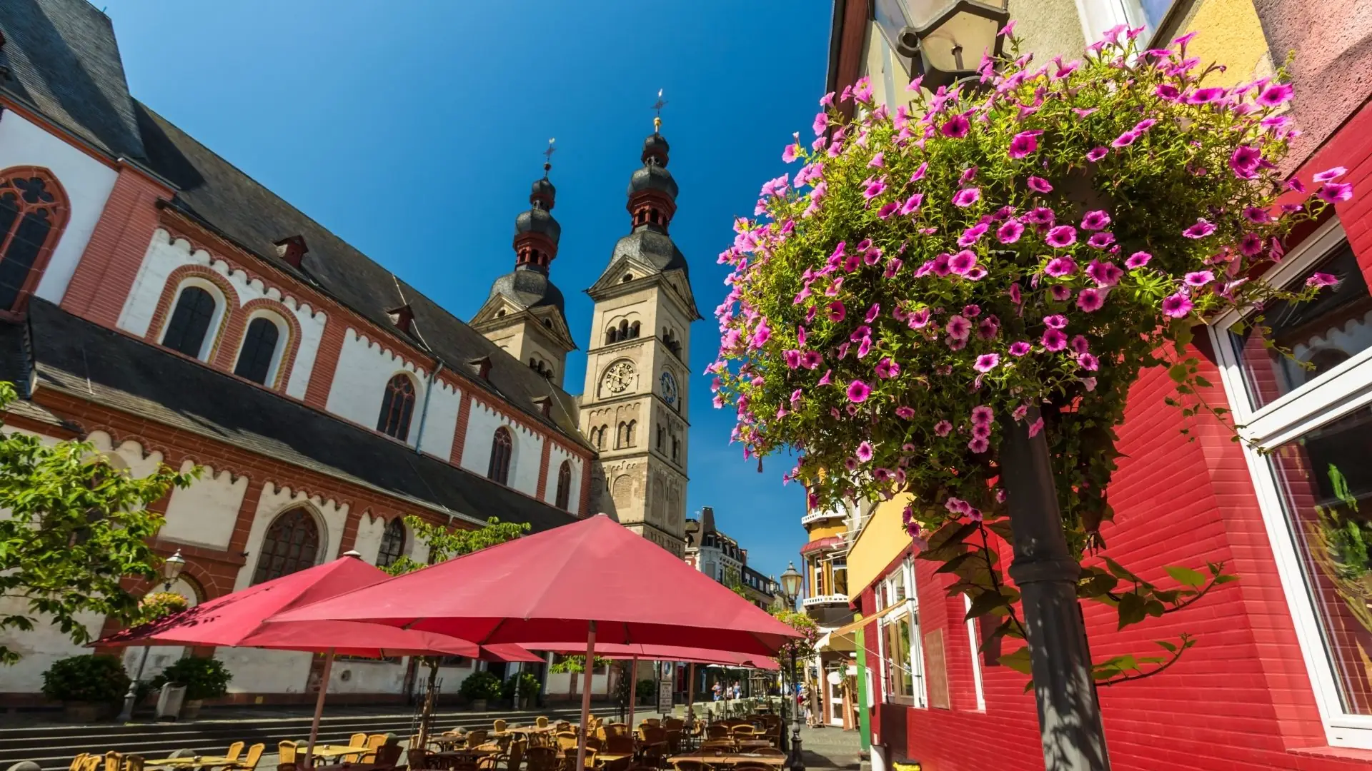Blick auf die Türme einer Kirche in der Altstadt von Koblenz mit blühenden Blumen und roten Sonnenschirmen eines Straßencafés
