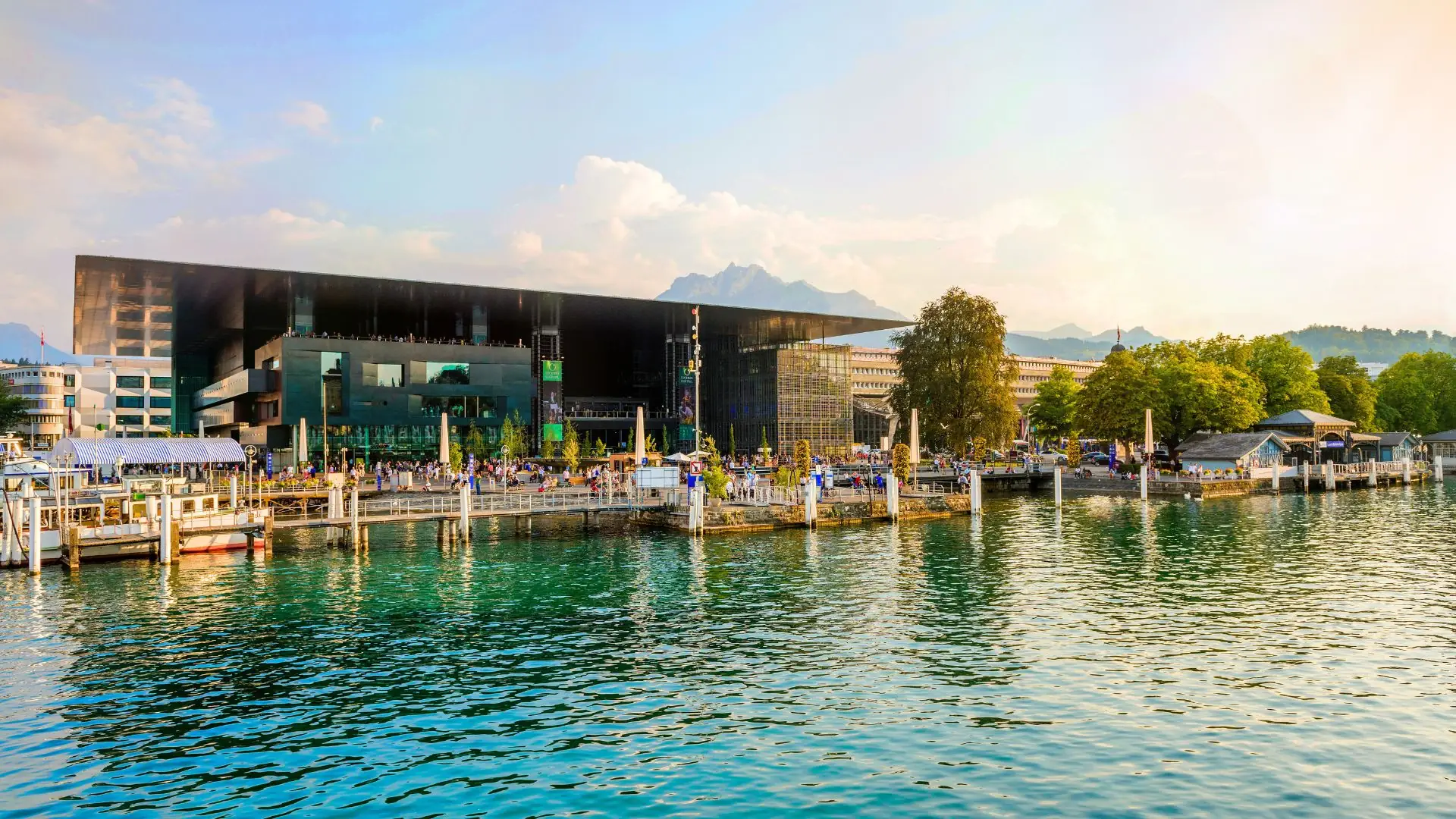 Das Kultur- und Kongresszentrum Luzern am Ufer des Vierwaldstättersees mit mehreren Bootsstegen und Bergen im Hintergrund.