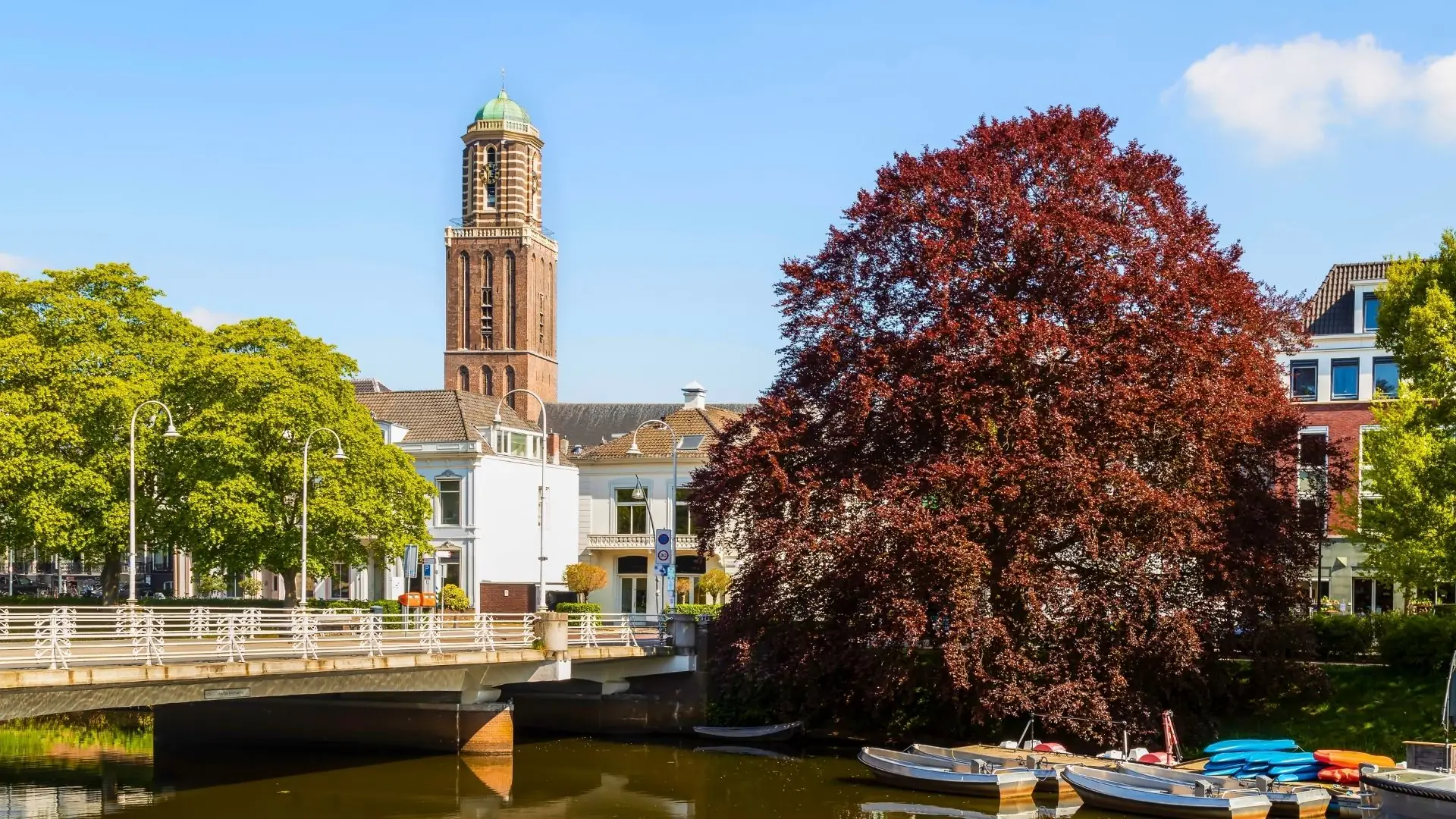 Liebfrauenbasilika Zwolle mit hohem Backsteinturm, davor Brücke über Kanal, rechts großer Baum mit roten Blättern, links grüne Bäume und Häuser.