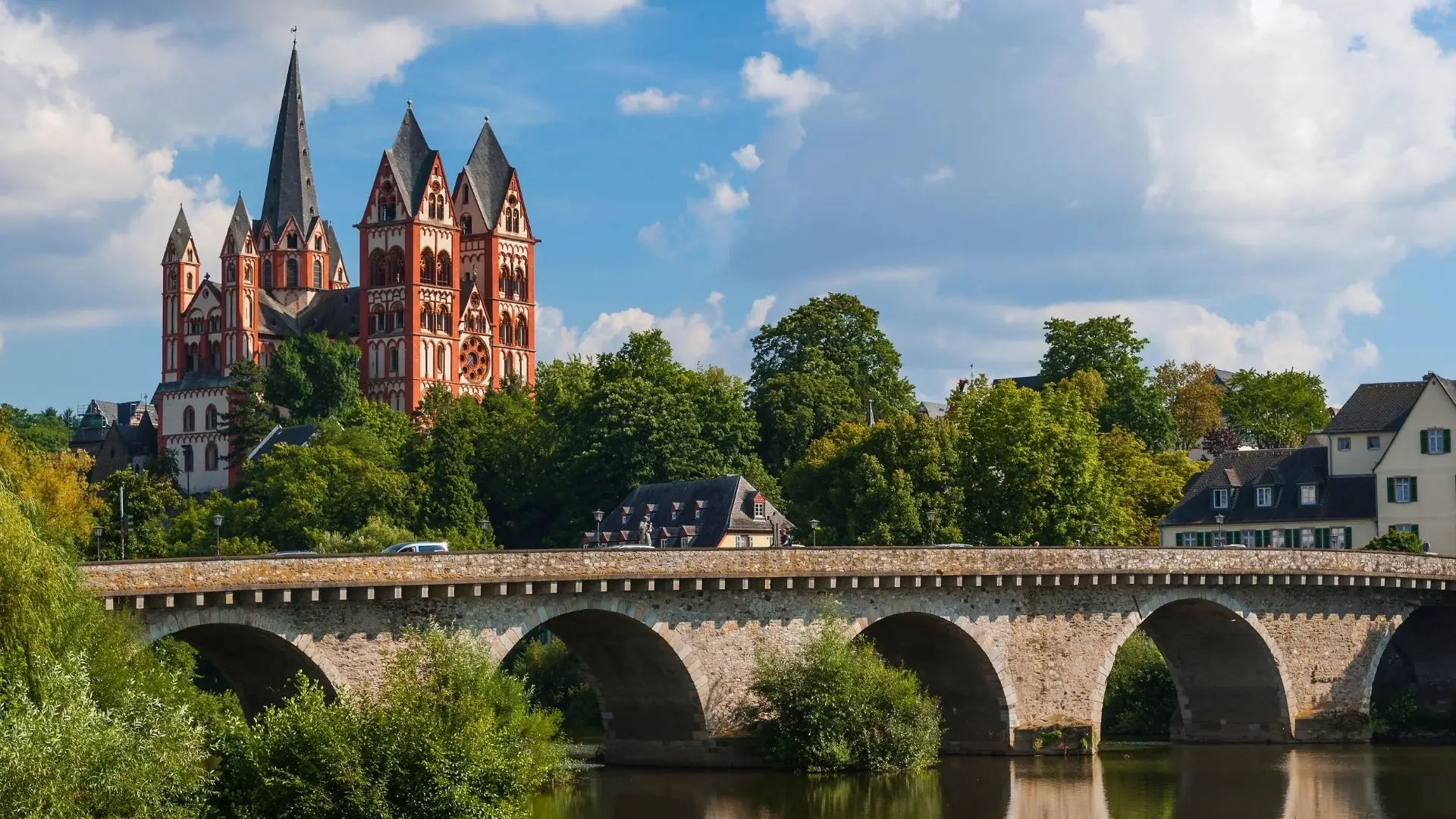 Gotische Kathedrale mit mehreren spitzen Türmen hinter einer Steinbrücke über einen Fluss, umgeben von Bäumen und Häusern.