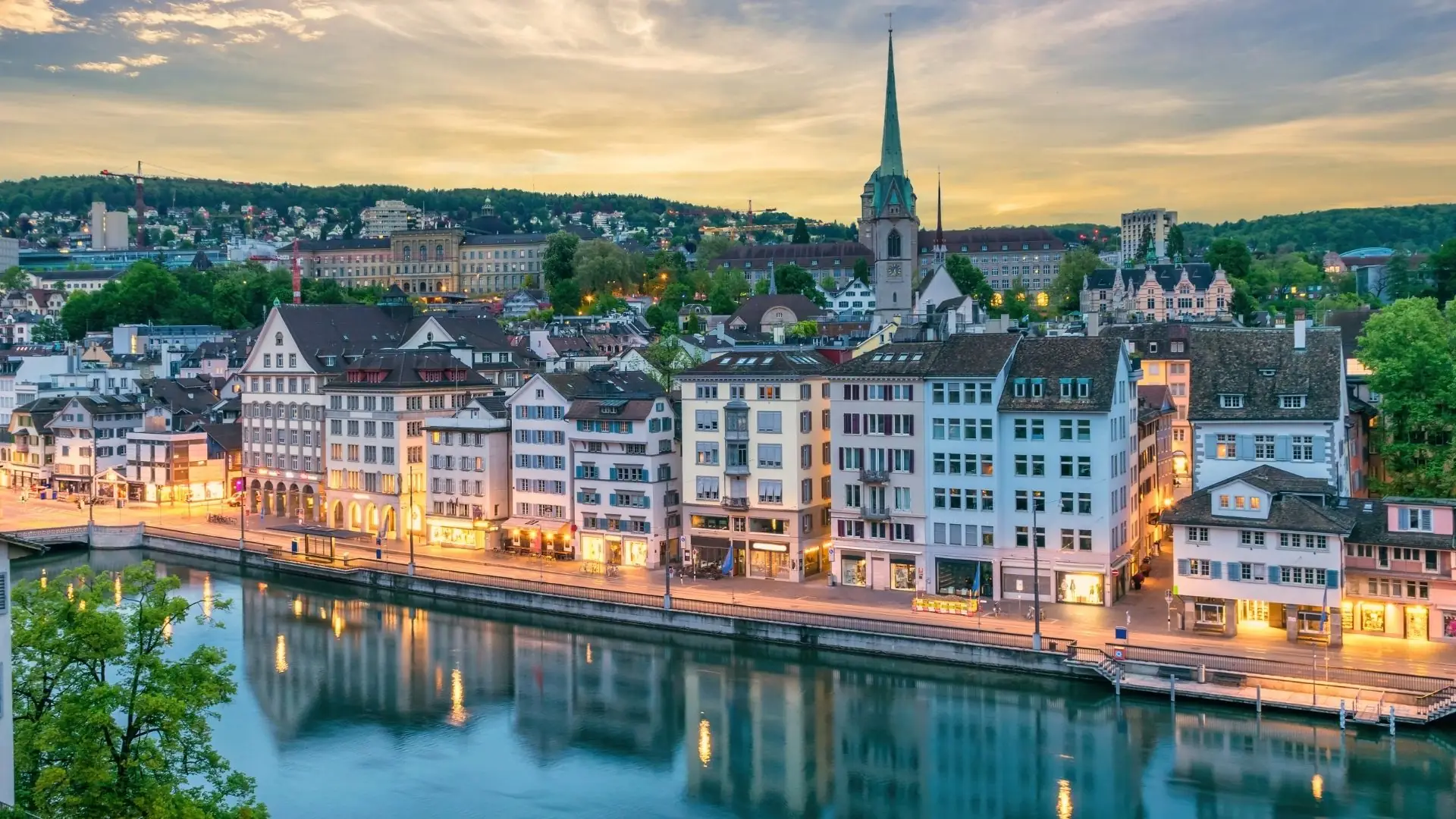 Blick auf die Häuserzeile am Ufer der Limmat in Zürich mit der Kirche St. Peter und dem Lindenhof-Hügel im Hintergrund bei Sonnenuntergang.