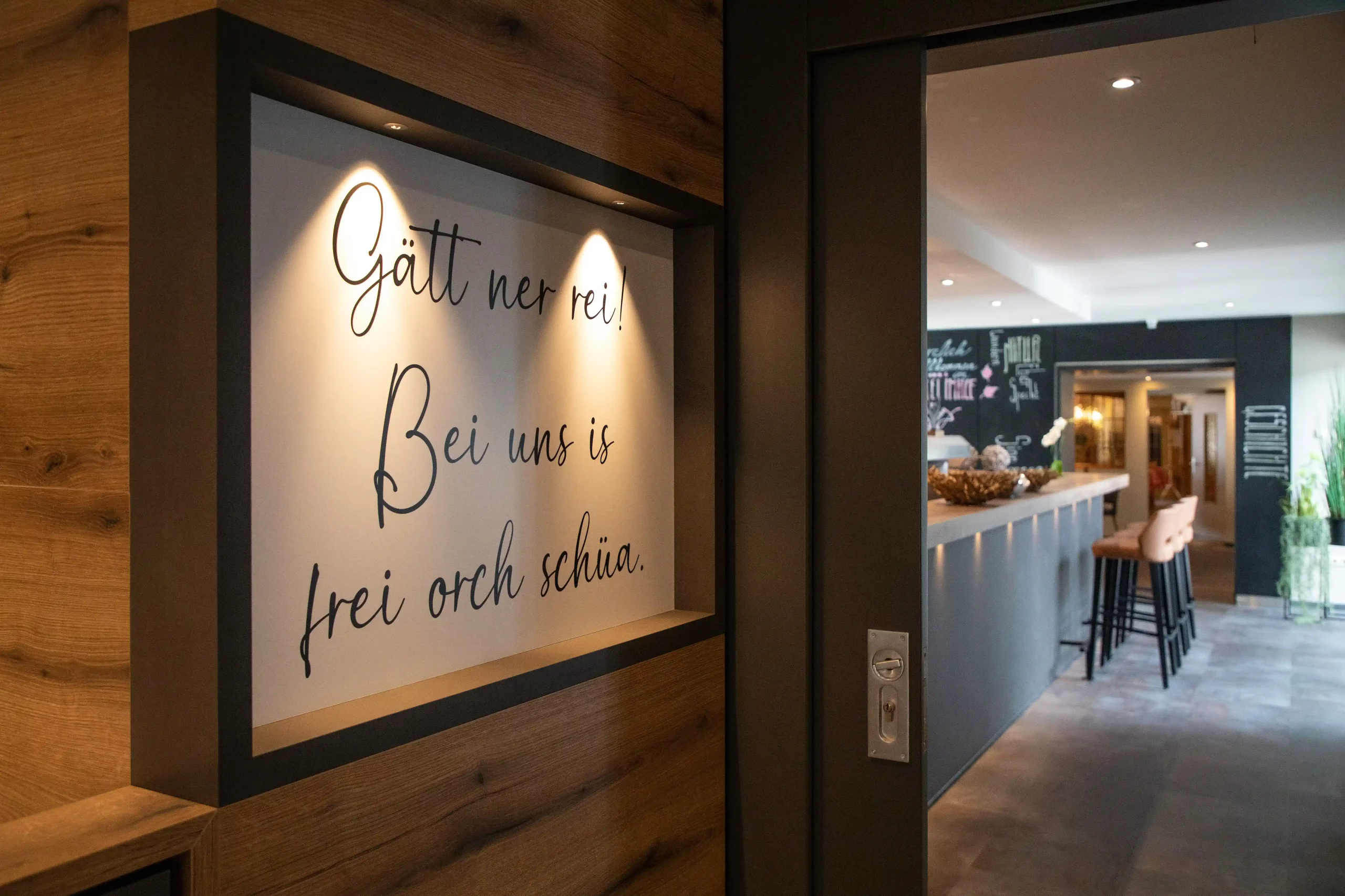 Beleuchtetes Schild mit der Aufschrift 'Gätt ner rei Bei uns is frei orch schüa' an einer Holzwand neben dem Eingang zur Bar im Hotel Imhof.