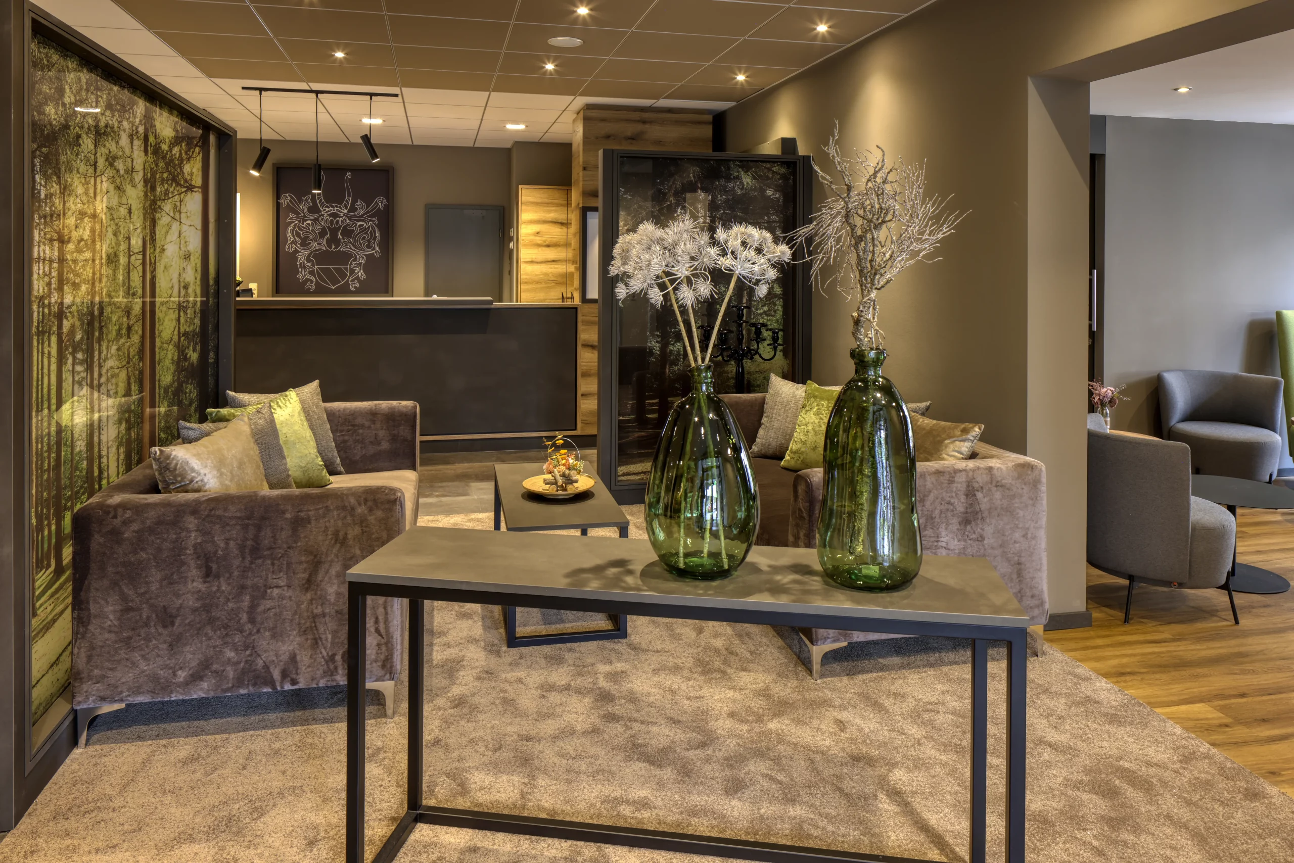 Moderne Hotellobby mit Rezeption im Hintergrund, zwei braune Sofas, grüne Vasen mit Trockenblumen auf einem Tisch und einem Teppich auf Holzboden.