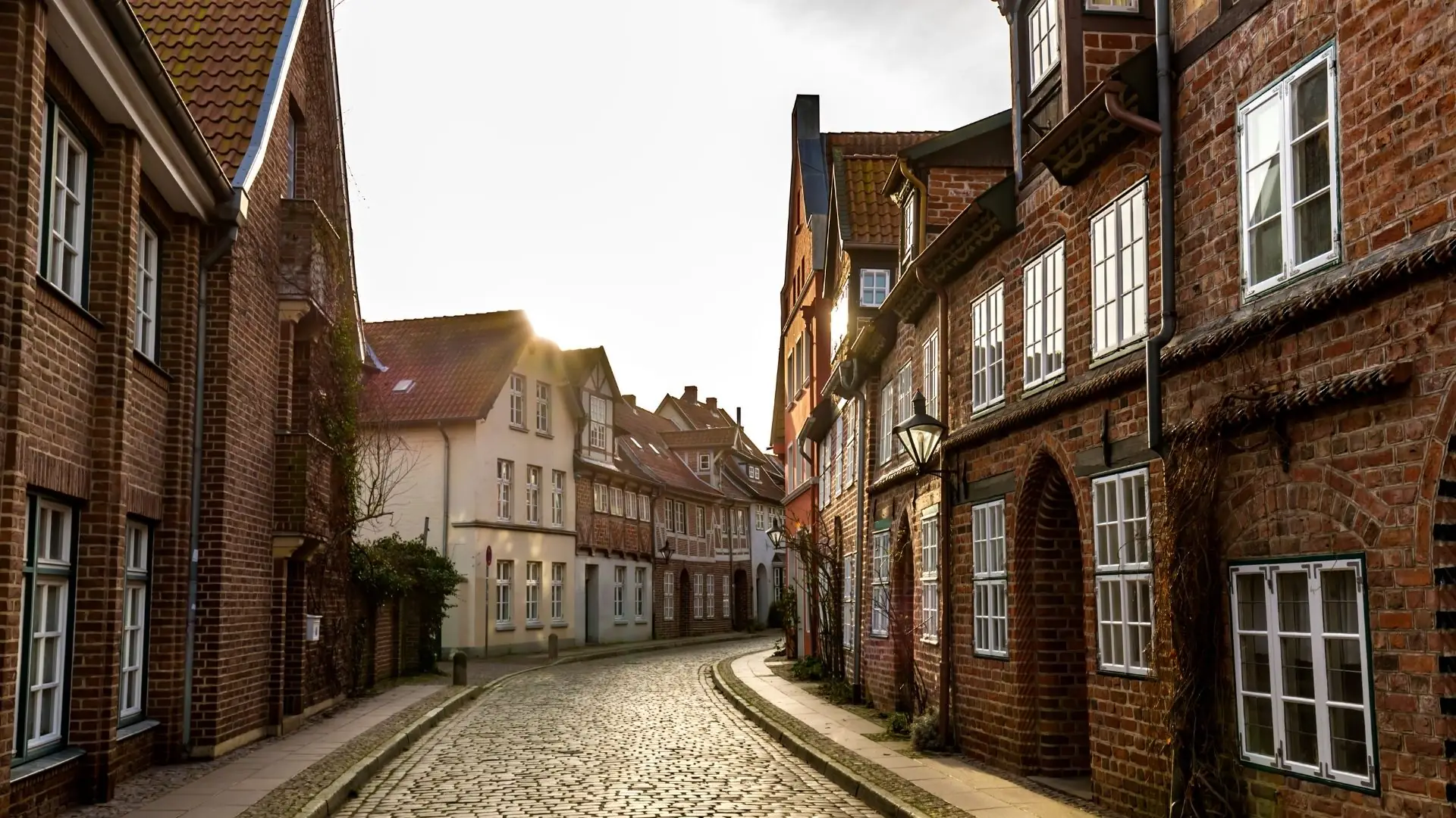 Enge Kopfsteinpflastergasse in Lüneburg mit historischen Backsteinhäusern und weißen Fenstern bei Sonnenuntergang.