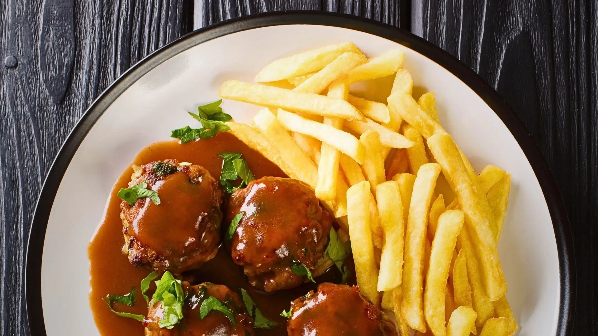 Teller mit zwei Boulets à la Liégeoise, übergossen mit brauner Sauce und mit gehackter Petersilie bestreut, daneben Pommes frites.