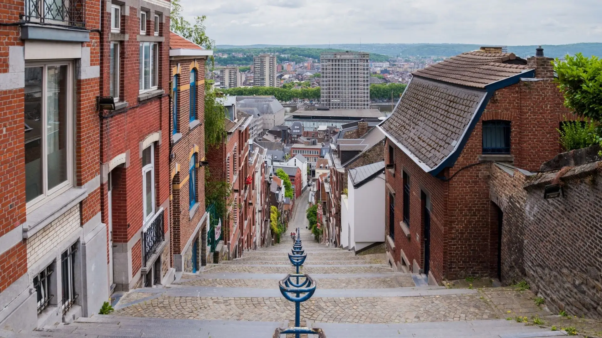 Blick die steile, gepflasterte Treppe in Lüttich hinab, flankiert von Backstein- und Putzfassaden, mit Stadt und Hügeln im Hintergrund.