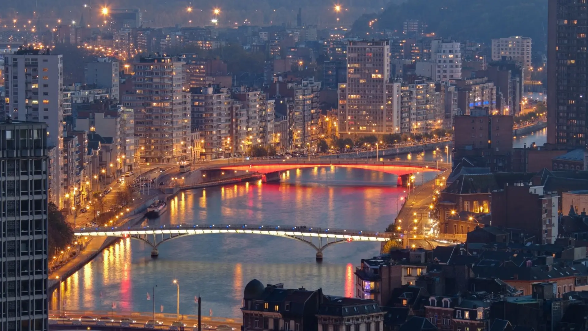 Nächtliche Stadtansicht von Lüttich mit beleuchteten Brücken über einen Fluss und Hochhäusern im Hintergrund.