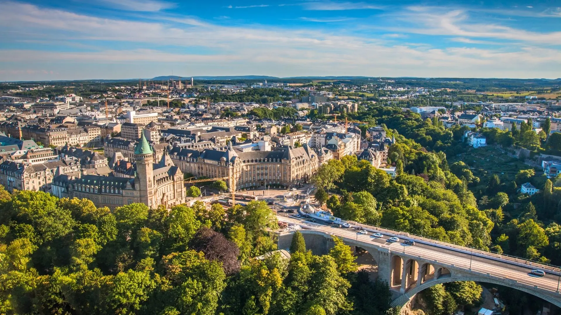 Luftaufnahme von Luxemburg mit dichtem Stadtzentrum, historischen Gebäuden, einer Brücke über ein bewaldetes Tal und blauem Himmel mit Wolken.
