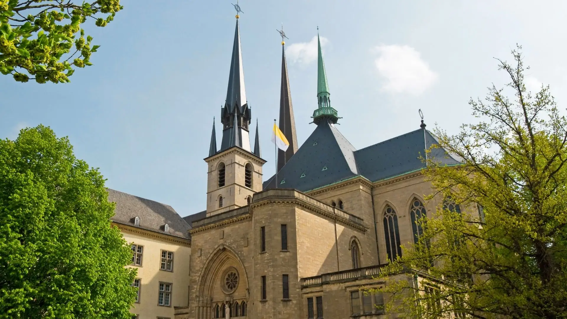 Gotische Notre-Dame-Kathedrale in Luxemburg mit drei hohen Türmen, spitzen Dächern und umgeben von grünen Bäumen
