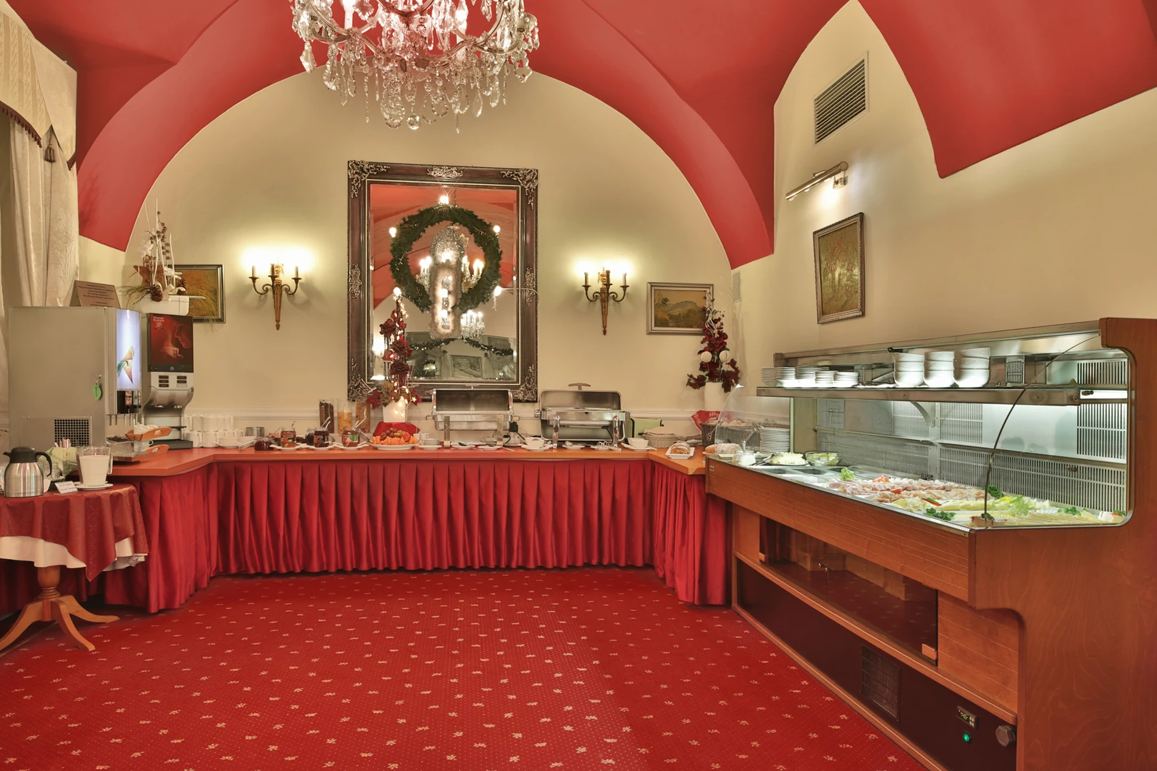 Das Bild zeigt einen eleganten Frühstücks- oder Buffetraum im Hotel Belvedere Marienbad, der mit luxuriösen Details und einer warmen Farbpalette gestaltet ist. Im Zentrum des Raumes dominiert ein beeindruckender Kristallleuchter, der eine gehobene und klassische Atmosphäre schafft. Die rote, geschwungene Decke in Kombination mit dem passend abgestimmten Teppichboden und der Tischverkleidung verleiht dem Raum eine opulente und einladende Ausstrahlung. Ein großes Buffet mit einer vielfältigen Auswahl an Speisen ist entlang der Wand arrangiert. Die Speisen sind auf sorgfältig arrangierten Platten und in Edelstahlbehältern präsentiert, ergänzt durch eine Kühlvitrine mit frischen Zutaten wie Salaten und Desserts. Der große Spiegel an der Wand, eingerahmt in einem kunstvoll verzierten Rahmen, reflektiert das Licht des Kronleuchters und lässt den Raum noch großzügiger wirken. Wandlampen und Dekorationselemente, wie festliche Kränze und Blumengestecke, runden das elegante Ambiente ab. Dieser Raum lädt Gäste ein, ihr Frühstück oder eine Mahlzeit in einem stilvollen, traditionellen Umfeld zu genießen und sich von der Aufmerksamkeit zum Detail begeistern zu lassen.
