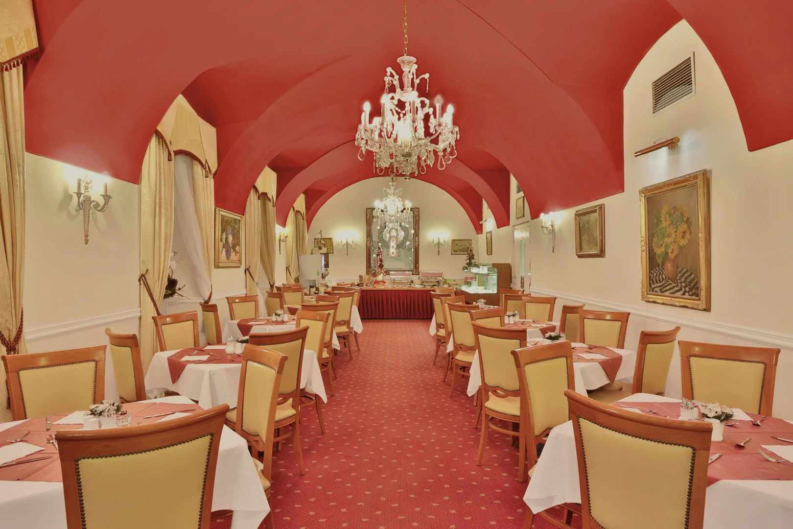 Das Bild zeigt den eleganten Speisesaal des Belvedere Marienbad, der mit einer Mischung aus klassischem Stil und luxuriöser Einrichtung beeindruckt. Der Raum ist geprägt von einer markanten roten Gewölbedecke, die in Kombination mit den warmen Farben des Teppichbodens und der Möbel eine einladende Atmosphäre schafft. Die langen Reihen stilvoll gedeckter Tische sind mit weißen Tischdecken und roten Akzenten dekoriert. Bequeme Holzstühle mit gepolsterten Rückenlehnen in Beige unterstreichen das gehobene Ambiente. Im Hintergrund zieht ein prächtiger Kristallleuchter die Aufmerksamkeit auf sich und verstärkt die luxuriöse Ausstrahlung des Raumes. Die Wände sind mit kunstvollen Gemälden und eleganten Wandlampen geschmückt, während die großen Fenster mit geschmackvollen Vorhängen ein zusätzliches Gefühl von Raum und Licht vermitteln. Der Buffetbereich am Ende des Raumes ist zentral positioniert und bietet eine großzügige Auswahl an Speisen, die in einer harmonischen Umgebung präsentiert werden. Dieser Speisesaal des Belvedere Marienbad vereint traditionelle Eleganz mit zeitloser Schönheit und bietet den perfekten Rahmen für ein gehobenes kulinarisches Erlebnis.