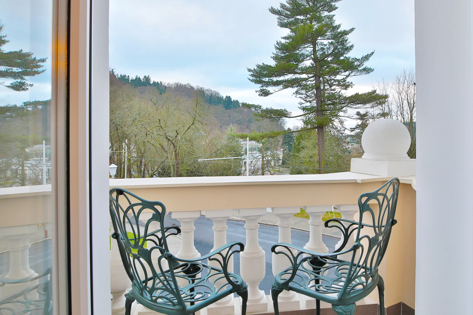 Das Bild zeigt den Balkon eines Zimmers im Hotel Belvedere Marienbad, der einen malerischen Blick auf die umgebende Natur bietet. Der Balkon ist mit zwei eleganten Metallstühlen im klassischen Design ausgestattet, die zu entspannten Momenten unter freiem Himmel einladen. Die Balustrade mit ihren weißen Säulen und dekorativen Details verleiht dem Außenbereich eine stilvolle und gehobene Atmosphäre. Im Hintergrund erstreckt sich eine idyllische Landschaft mit hohen, grünen Bäumen, die die Ruhe und Schönheit der Umgebung unterstreichen. Die sanfte Hügellandschaft und die klaren Linien des Himmels sorgen für ein harmonisches Gesamtbild und laden dazu ein, die Natur und frische Luft zu genießen. Dieser Balkon bietet den perfekten Ort, um den Tag mit einem Kaffee zu beginnen oder den Abend entspannt ausklingen zu lassen – ein weiteres Highlight, das den Charme des Belvedere Marienbad unterstreicht.