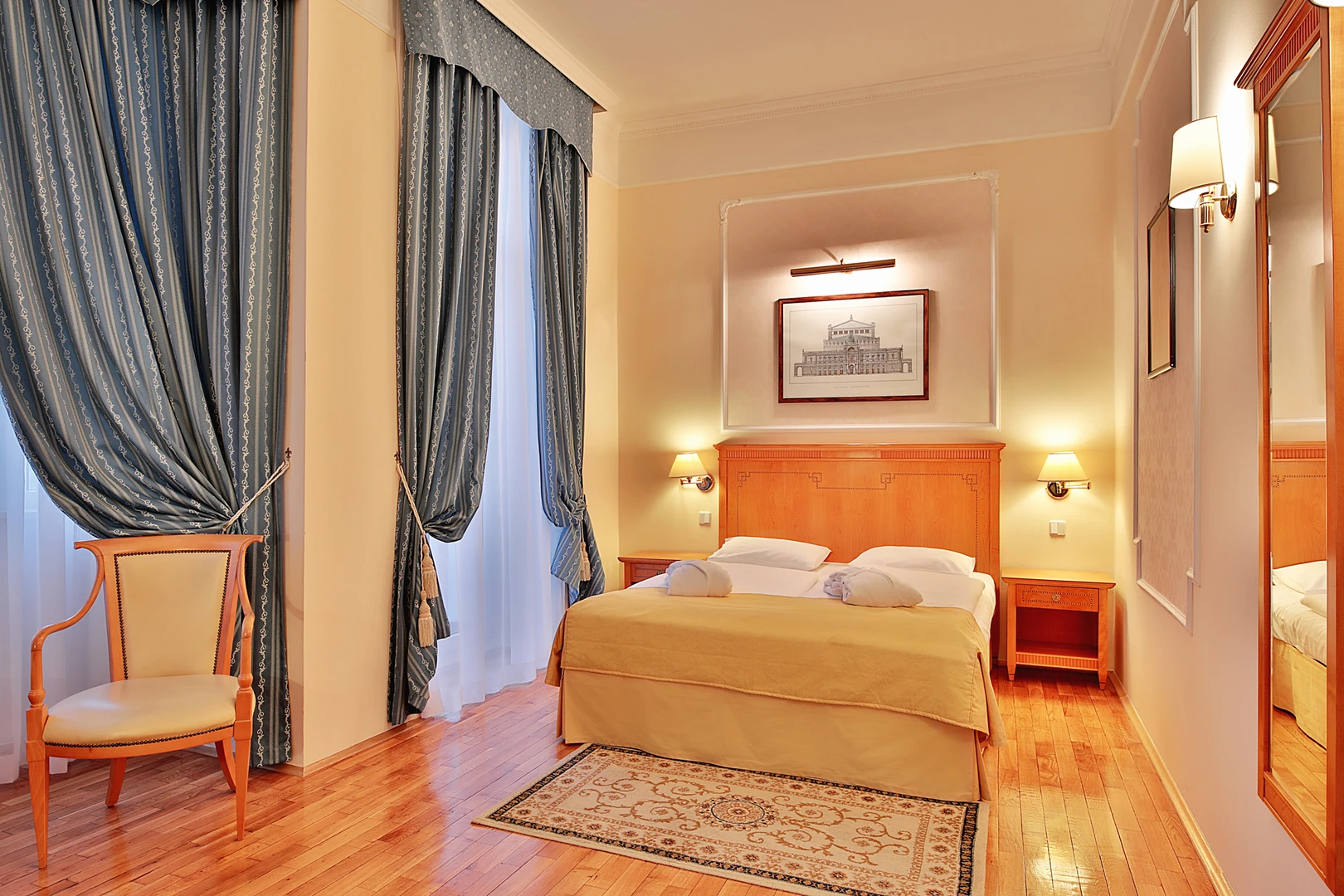 Das Bild zeigt ein stilvolles Gästezimmer im Hotel Belvedere Marienbad, das durch seine warme und einladende Atmosphäre besticht. Im Mittelpunkt steht ein elegantes Doppelbett mit einem hochwertigen Holzkopfteil, ergänzt durch weiche Bettwäsche und sorgfältig gefaltete Handtücher, die für einen einladenden ersten Eindruck sorgen. Die großen Fenster sind mit schweren Vorhängen in einem edlen Blau dekoriert, die durch dezente Muster und elegante Drapierungen den Raum harmonisch ergänzen. Der helle Holzboden und ein kleiner, kunstvoll gemusterter Teppich verleihen dem Zimmer eine angenehme Wärme und unterstreichen das klassische Design. Ein gepolsterter Stuhl mit Holzrahmen steht in einer gemütlichen Ecke, ideal zum Lesen oder Entspannen. Die Wände sind in zarten Beigetönen gehalten und mit einem großen Spiegel sowie kunstvollen Wandverzierungen dekoriert, die den Raum optisch erweitern und ihm ein gehobenes Ambiente verleihen. Das Zimmer verbindet traditionelle Eleganz mit modernem Komfort und spiegelt den hochwertigen Standard des Hotels wider – perfekt für einen entspannten und erholsamen Aufenthalt in Marienbad.