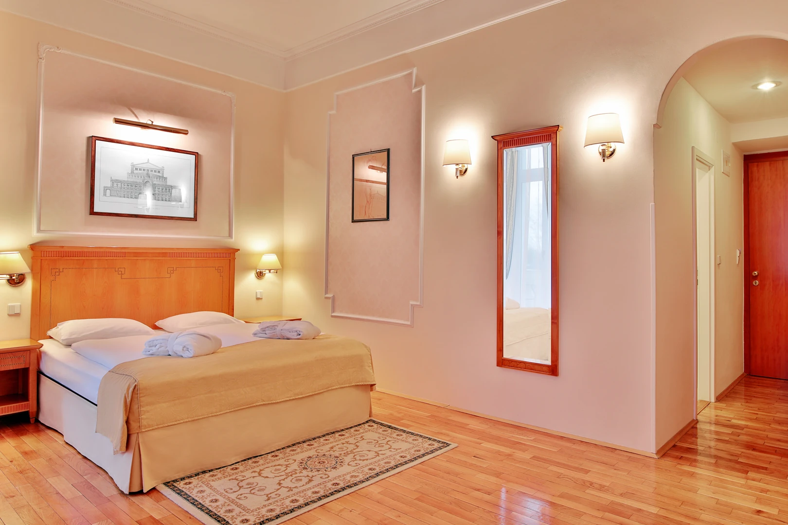 Das Bild zeigt ein geschmackvoll eingerichtetes Gästezimmer im Hotel Belvedere Marienbad, das Komfort und Eleganz harmonisch verbindet. Im Zentrum des Raumes steht ein großes Doppelbett mit einem hochwertigen Holzrahmen und einer weichen Bettwäsche in warmen Beige- und Cremetönen. Auf dem Bett sind sorgfältig gefaltete Handtücher mit Bademänteln platziert, was den Gästen einen einladenden Empfang bereitet. Der Holzboden und ein eleganter, gemusterter Teppich verleihen dem Raum eine warme und gemütliche Atmosphäre. Die Wände sind in sanften Pastellfarben gehalten und mit dezenten Stuckelementen sowie geschmackvollen Kunstwerken geschmückt, die eine klassische Ästhetik betonen. Ein großer Wandspiegel mit einem Holzrahmen reflektiert das Licht und verstärkt das Gefühl von Helligkeit und Raum. Sanftes Licht von den stilvollen Wandlampen schafft eine entspannende und angenehme Beleuchtung, während der Durchgang zur Tür mit einem sanften Rundbogen eine charmante architektonische Note hinzufügt. Dieses Zimmer bietet eine ideale Mischung aus Funktionalität und Stil, die den hohen Standard des Belvedere Marienbad widerspiegelt und einen angenehmen Aufenthalt verspricht.