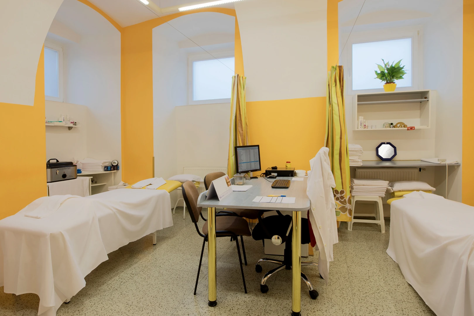 Das Bild zeigt einen hellen und einladenden Behandlungsraum im Hotel Belvedere Marienbad, der für professionelle Wellness- oder medizinische Anwendungen gestaltet wurde. Der Raum ist in einer freundlichen Gelb-Weiß-Farbkombination gehalten, die eine warme und beruhigende Atmosphäre schafft. Im Raum befinden sich mehrere Behandlungsliegen, die mit weißen Tüchern bedeckt sind und für verschiedene Anwendungen vorbereitet wurden. Jede Liege bietet Privatsphäre, da sie durch Vorhänge abgetrennt werden kann. Ein zentral platzierter Schreibtisch mit einem Computer und Bürostühlen deutet darauf hin, dass der Raum auch für Konsultationen oder organisatorische Zwecke genutzt wird. An der Seite stehen Regale, die mit ordentlich gestapelten Handtüchern, Pflegeprodukten und medizinischen Utensilien ausgestattet sind. Eine kleine Pflanze in einem gelben Topf auf einem Regal fügt einen Hauch von Natur hinzu und verstärkt das einladende Ambiente. Der Raum ist funktional und zugleich angenehm gestaltet, um sowohl Komfort als auch Effizienz zu gewährleisten, und bietet Gästen des Belvedere Marienbad eine professionelle Umgebung für ihre Behandlungs- und Wellnessbedürfnisse.