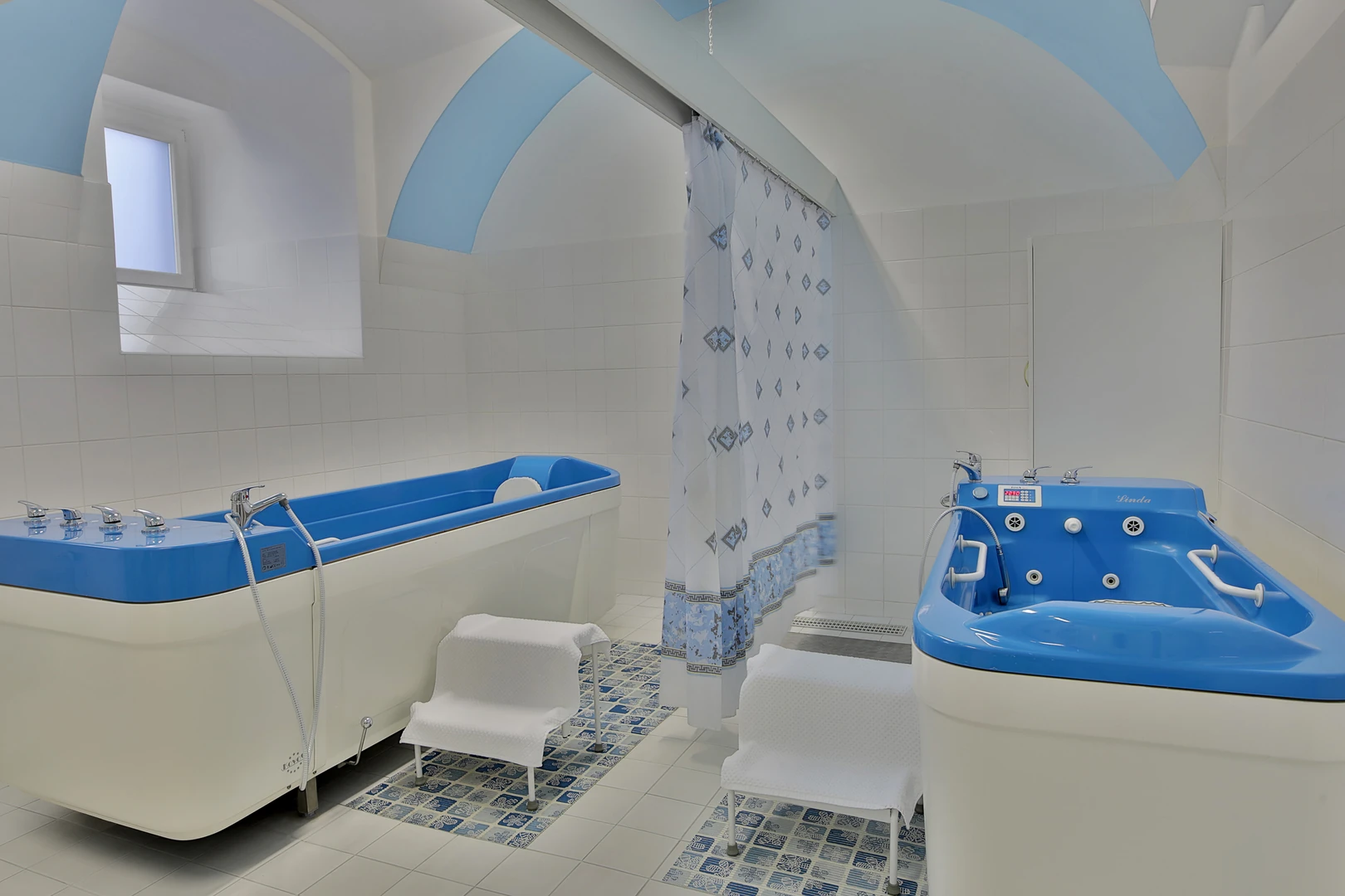 Das Bild zeigt einen modernen und professionell ausgestatteten Balneotherapieraum im Hotel Belvedere Marienbad. Der Raum ist hell und sauber gestaltet, mit weißen Wandfliesen und akzentuierten, hellblauen Bögen an der Decke, die eine beruhigende Atmosphäre schaffen. Im Mittelpunkt stehen zwei speziell ausgestattete Hydrotherapiewannen in einem auffälligen Blau-Weiß-Design. Diese Wannen sind für therapeutische Behandlungen konzipiert und mit modernen Armaturen sowie integrierten Massagedüsen ausgestattet. Die Bedienfelder und Haltegriffe an den Wannen garantieren eine sichere und komfortable Nutzung. Der Boden ist mit gemusterten Fliesen gestaltet, die zum harmonischen Farbschema des Raums passen. Kleine, mit Handtüchern bedeckte Stufen erleichtern den Zugang zu den Wannen und betonen den Komfort, der den Gästen geboten wird. Ein Duschvorhang trennt den Bereich für zusätzliche Privatsphäre. Dieser Raum kombiniert moderne Technologie mit elegantem Design und unterstreicht die hochwertige Ausstattung des Hotels Belvedere Marienbad, das seinen Gästen erstklassige Wellness- und Therapieanwendungen bietet.