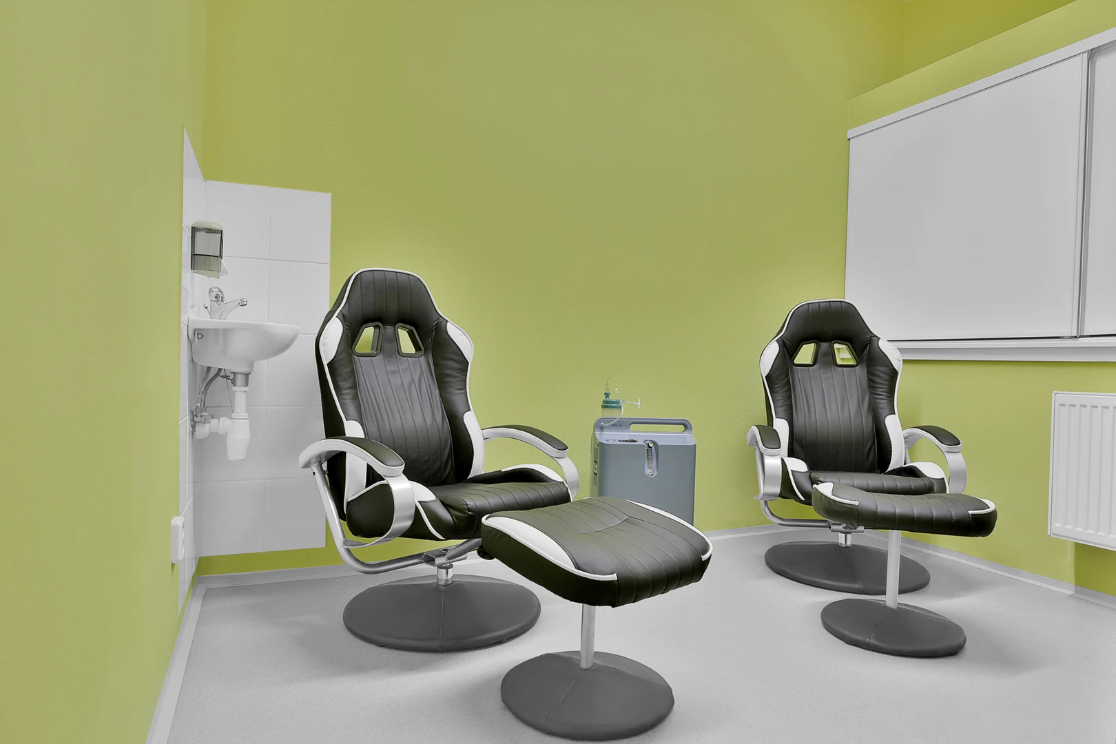 Das Bild zeigt einen modernen und funktional eingerichteten Behandlungsraum im Hotel Belvedere Marienbad, der für spezielle Wellness- oder Therapieanwendungen konzipiert ist. Der Raum ist in einem angenehmen, beruhigenden Grünton gestrichen, der eine entspannte Atmosphäre schafft und Harmonie ausstrahlt. Im Fokus stehen zwei ergonomische, gepolsterte Stühle in einem stilvollen Schwarz-Weiß-Design. Die Stühle sind mit passenden Fußhockern ausgestattet, die maximalen Komfort für Anwendungen oder Entspannungsphasen bieten. Neben den Stühlen ist ein medizinisches Gerät platziert, das auf spezielle Behandlungsoptionen wie Sauerstofftherapien oder ähnliche Anwendungen hinweist. Ein kleines Waschbecken an der Seite unterstreicht die praktische und hygienische Ausstattung des Raumes, während ein schlichtes, modernes Design im gesamten Raum beibehalten wird. Der Raum ist klar strukturiert und bietet Gästen eine komfortable Umgebung für personalisierte Wellness- und Therapieerfahrungen.