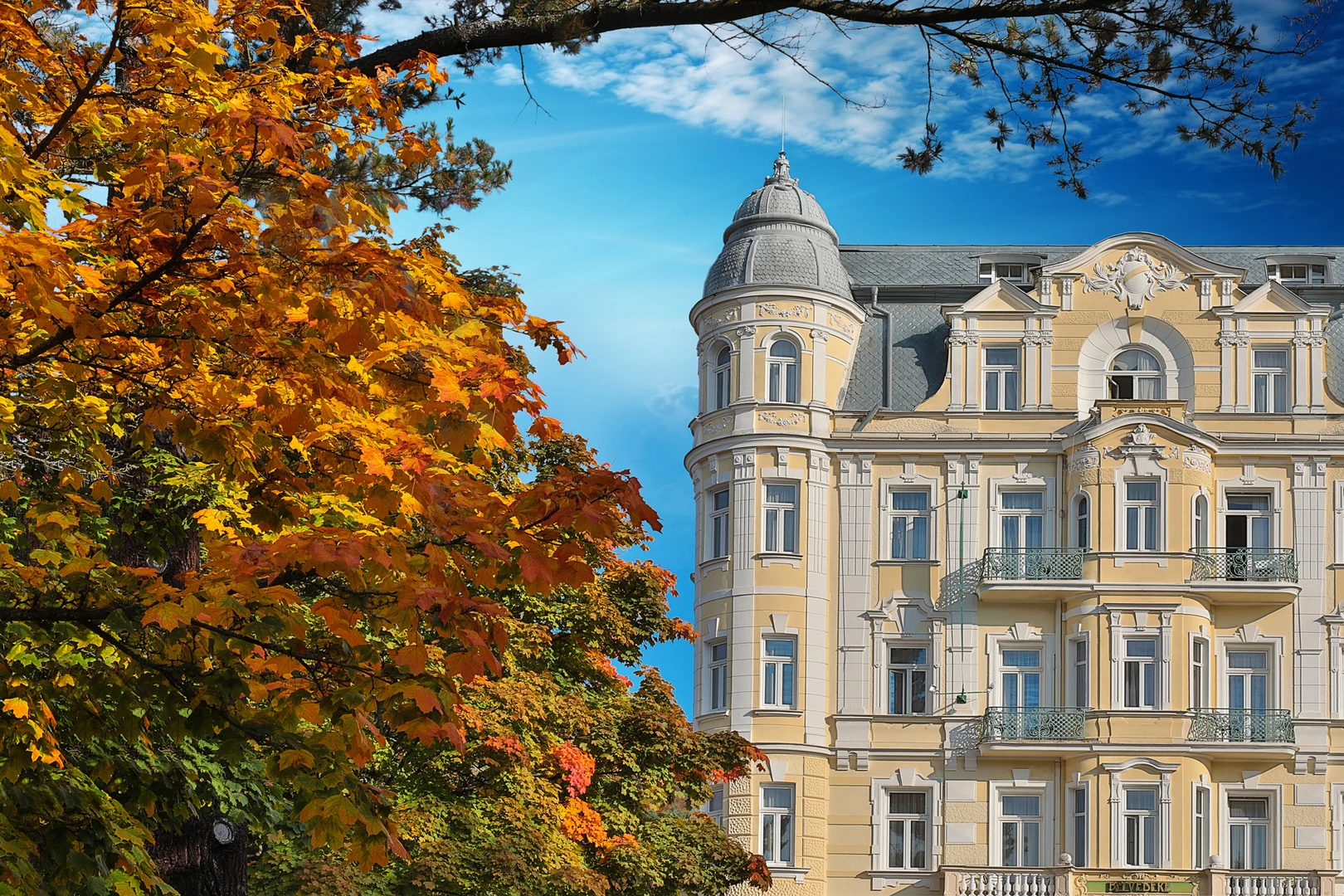 Das Bild zeigt eine malerische Ansicht des Hotel Belvedere Marienbad im herbstlichen Ambiente. Die historische Fassade des Hotels beeindruckt mit ihrer eleganten Architektur im Jugendstil, gekennzeichnet durch zarte Gelbtöne, kunstvolle Verzierungen und geschwungene Balkone mit filigranen Geländern. Besonders hervorzuheben ist der runde Turm mit seiner dekorativen Kuppel, die dem Gebäude einen majestätischen Charakter verleiht. Im Vordergrund leuchten die herbstlichen Blätter eines Baumes in satten Gelb- und Orangetönen, die eine warme und einladende Atmosphäre schaffen und einen reizvollen Kontrast zum klaren blauen Himmel bilden. Die Kombination aus der prächtigen Natur und der kunstvollen Architektur vermittelt das einzigartige Flair von Marienbad als Ort von Eleganz und Ruhe. Dieses Bild fängt perfekt den historischen Charme und die natürliche Schönheit des Hotels Belvedere und seiner Umgebung ein, was es zu einer idealen Destination für Erholung und Genuss macht.